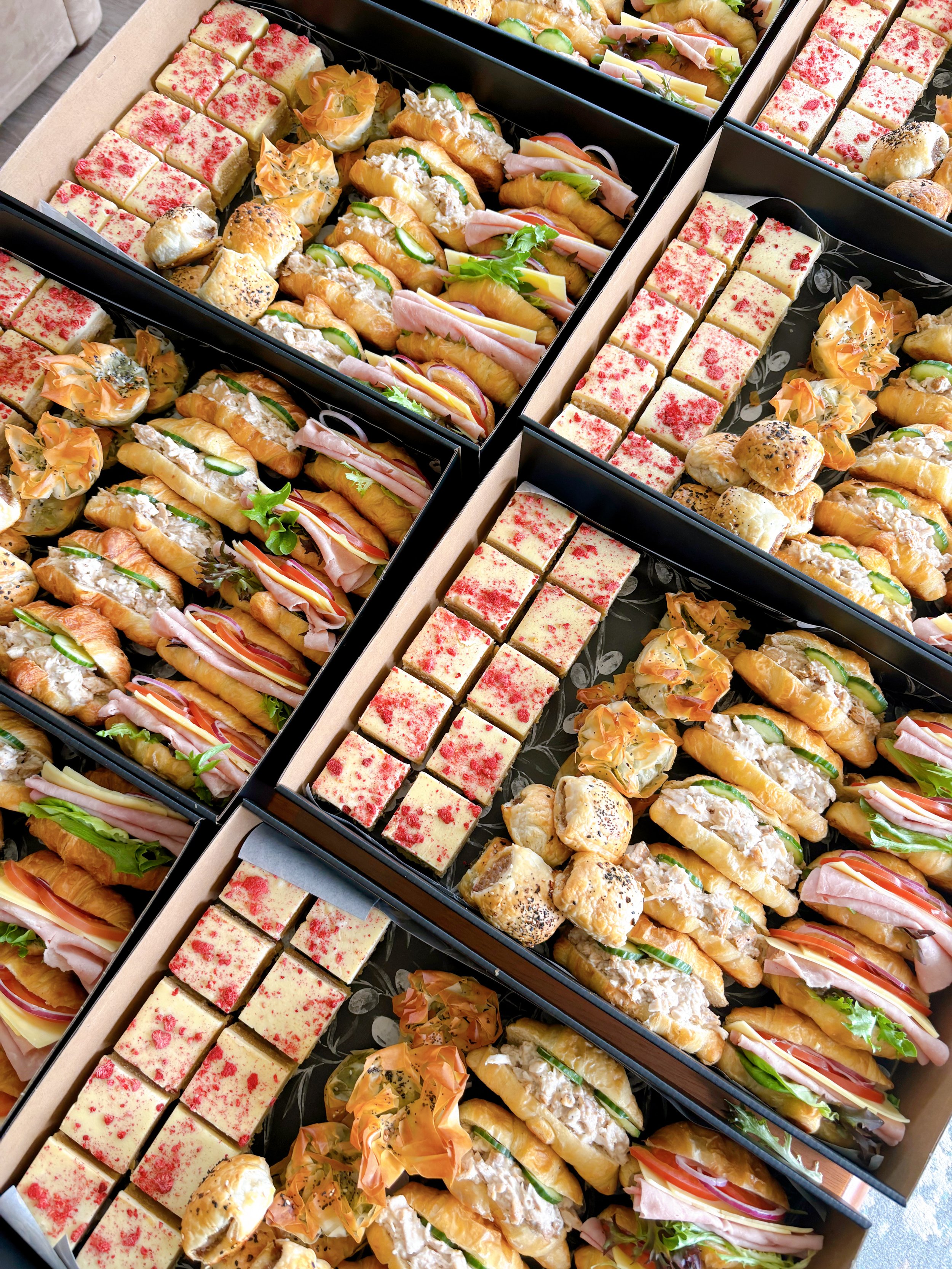 Assorted sandwiches, including croissants and small cake squares with red topping, arranged in open black boxes.