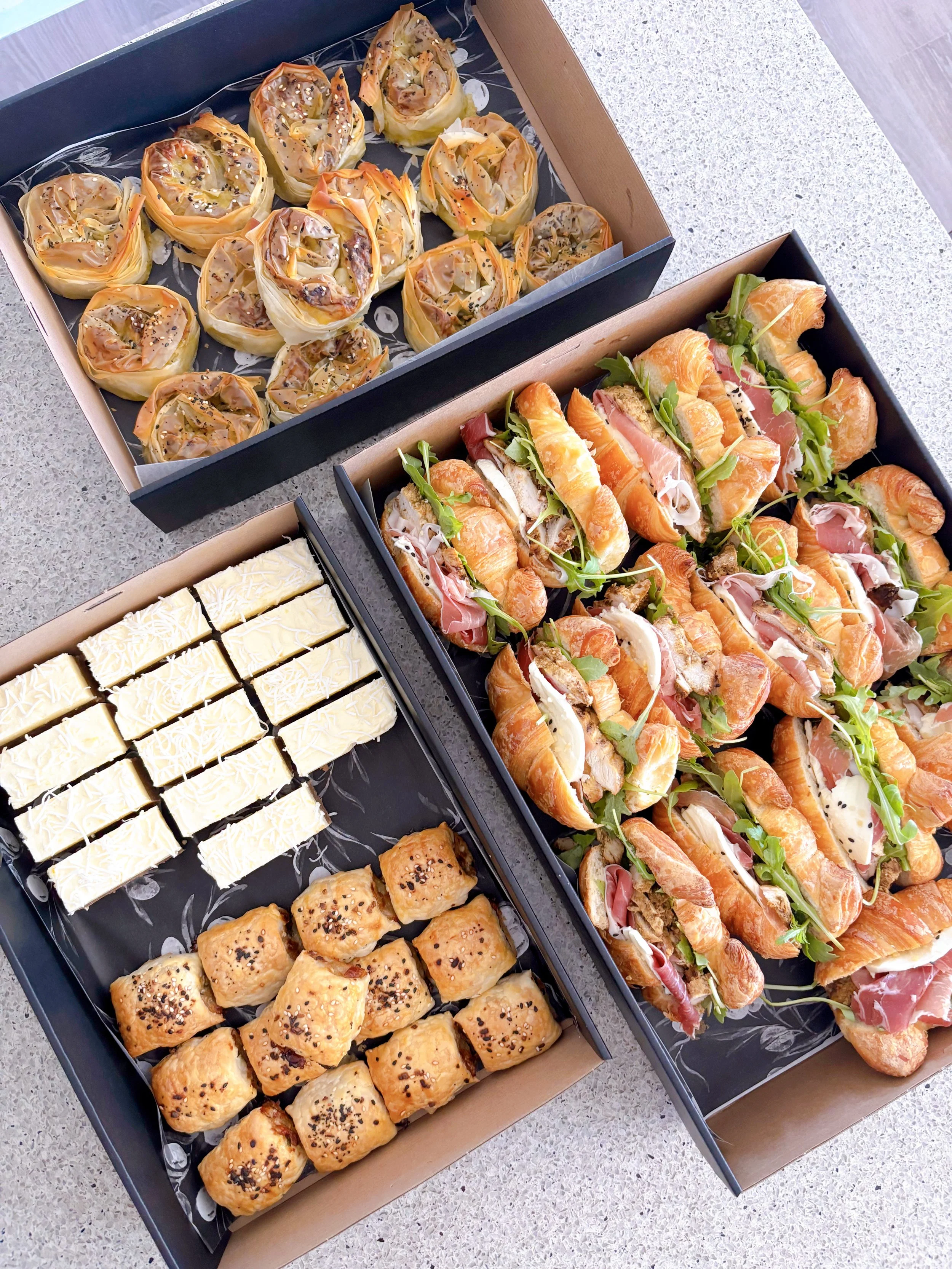 Assorted pastries, sandwiches, and desserts in open boxes on a kitchen counter.