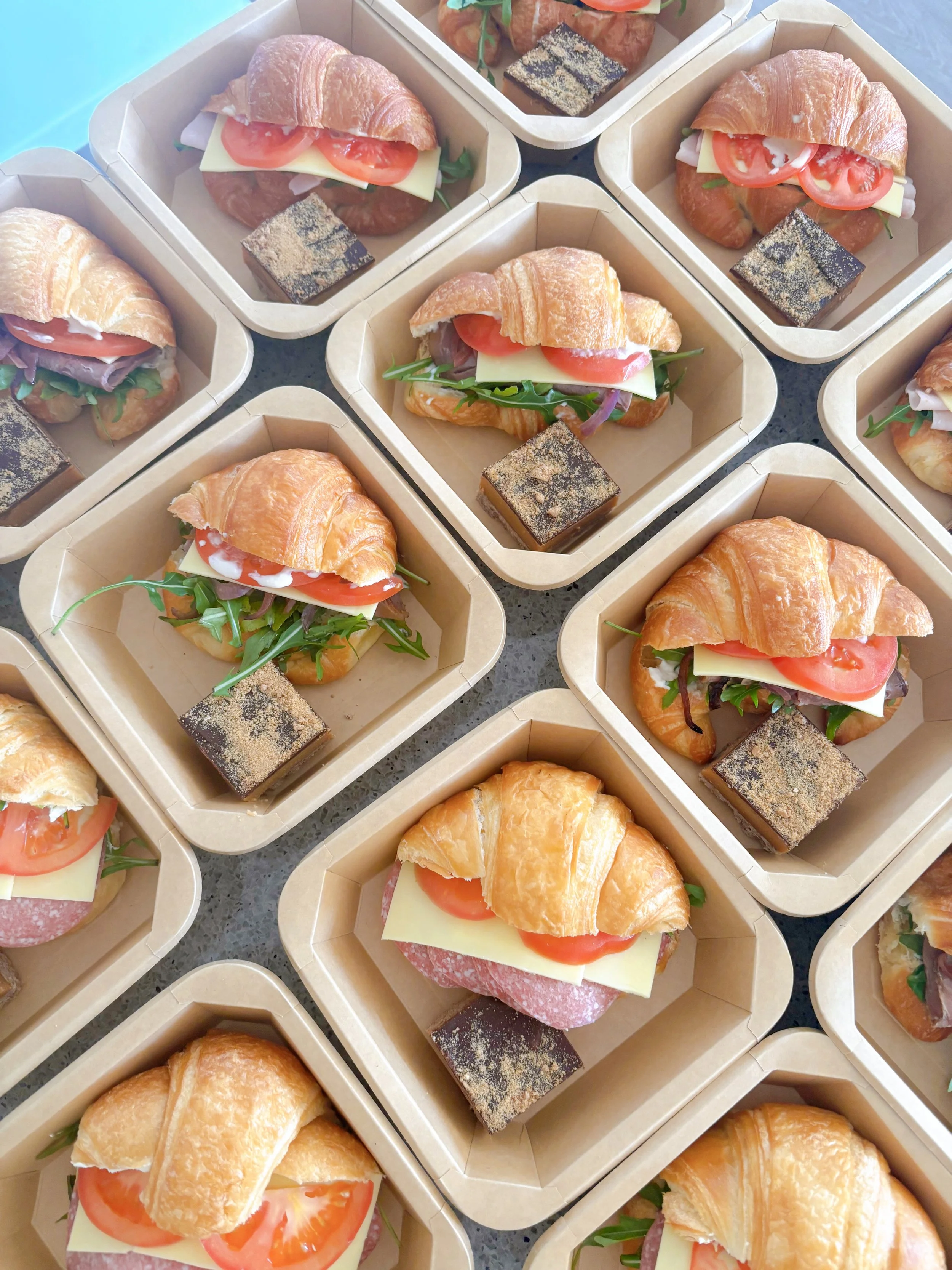 Multiple takeaway boxes containing croissant sandwiches with tomato, cheese, and greens, along with small square chocolate desserts.