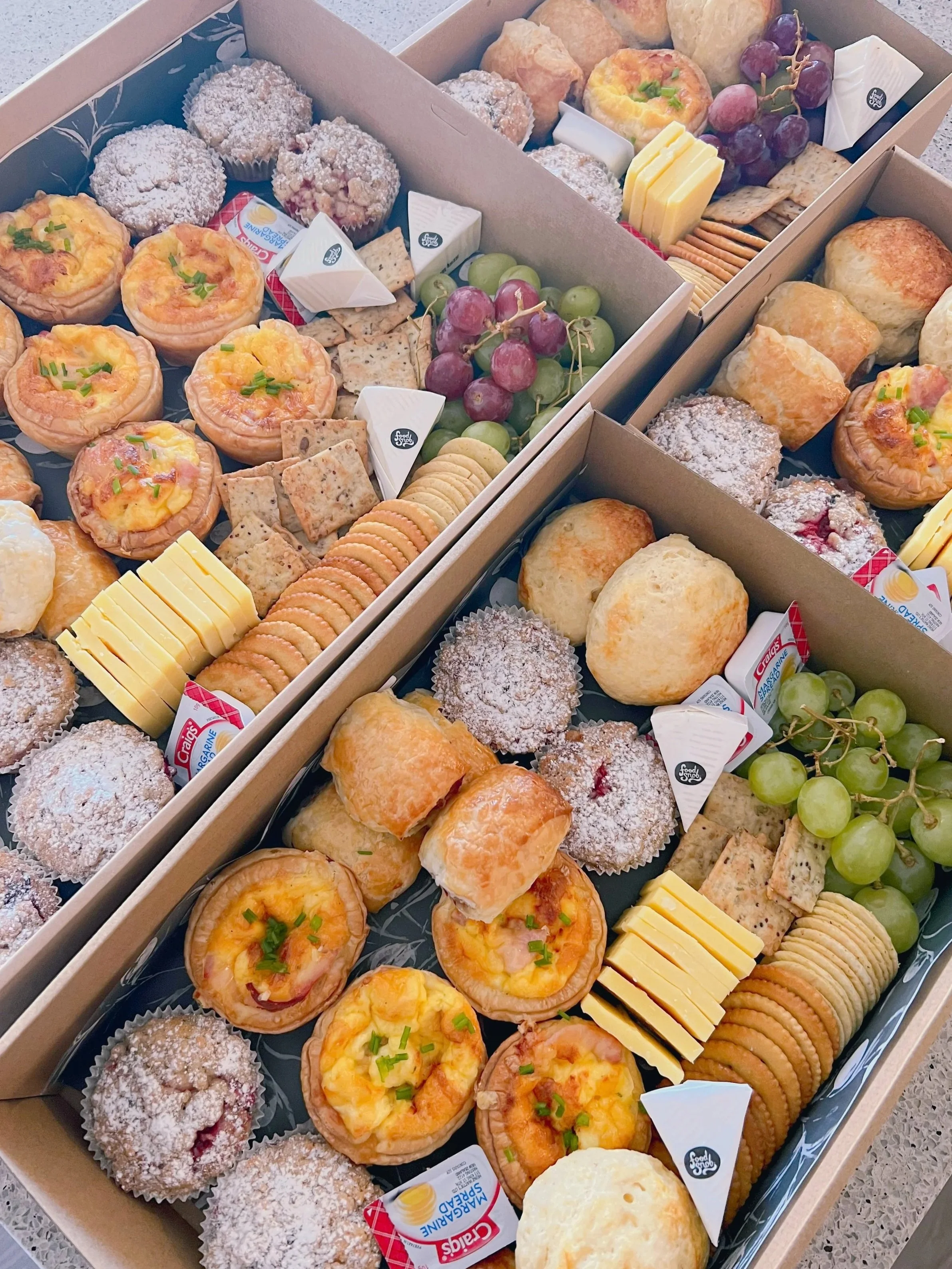 Trays of assorted pastries, including mini muffins, cheese and fruit tarts, crackers, grapes, sliced cheese, biscuits, and flavored spreads.