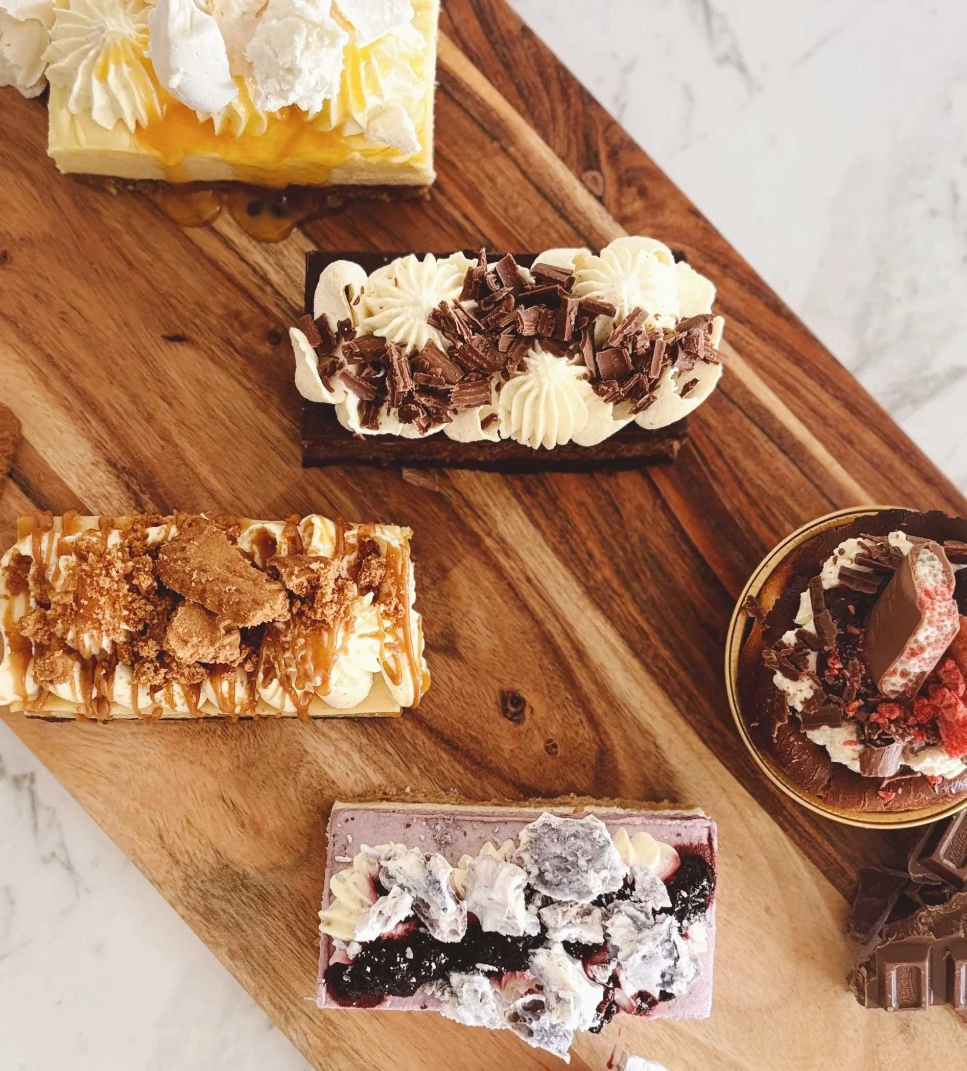 Assorted decorated desserts on a wooden surface, including a lemon-flavored cake, a chocolate-topped cream cake, a caramel and cookie-topped pastry, a berry and cream cake, and a cup of chocolate mousse with toppings.