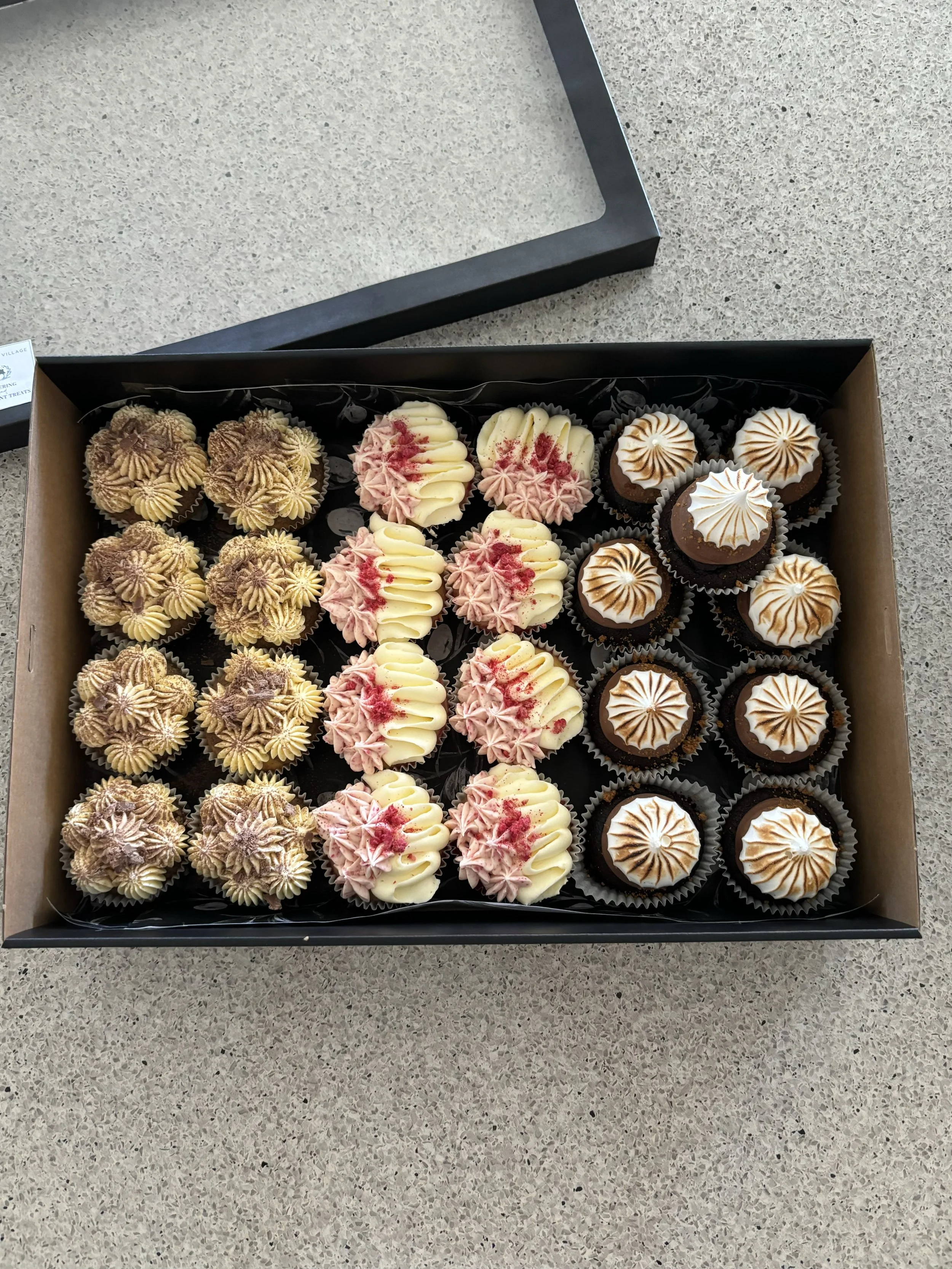 A box of assorted cupcakes with three different styles of frosting, on a gray countertop.
