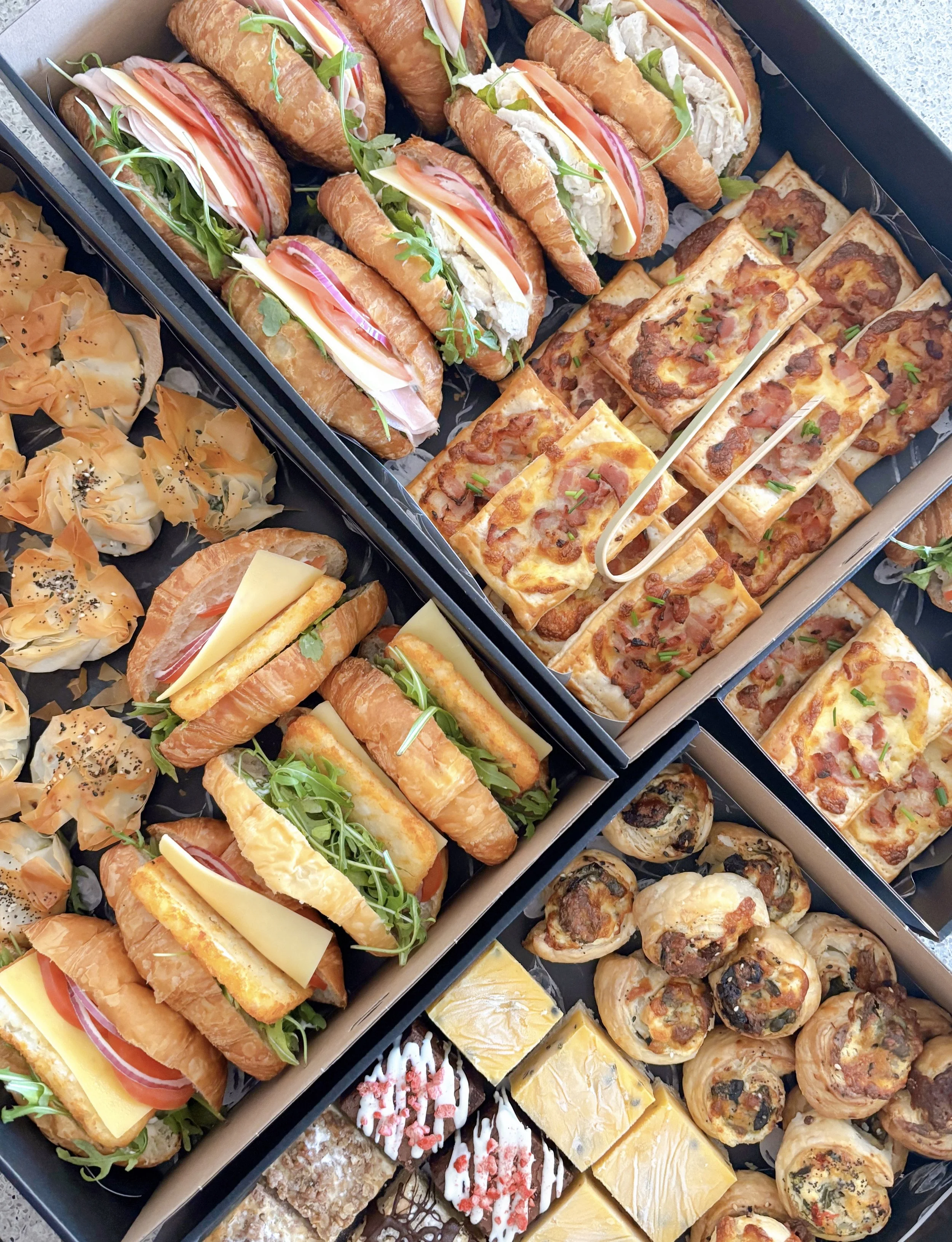 A variety of party foods including mini sandwiches with meats, cheeses, and greens, flatbread pizzas with tomato sauce and cheese, small savory pastries, and assorted desserts such as cake squares and rolled pastries.