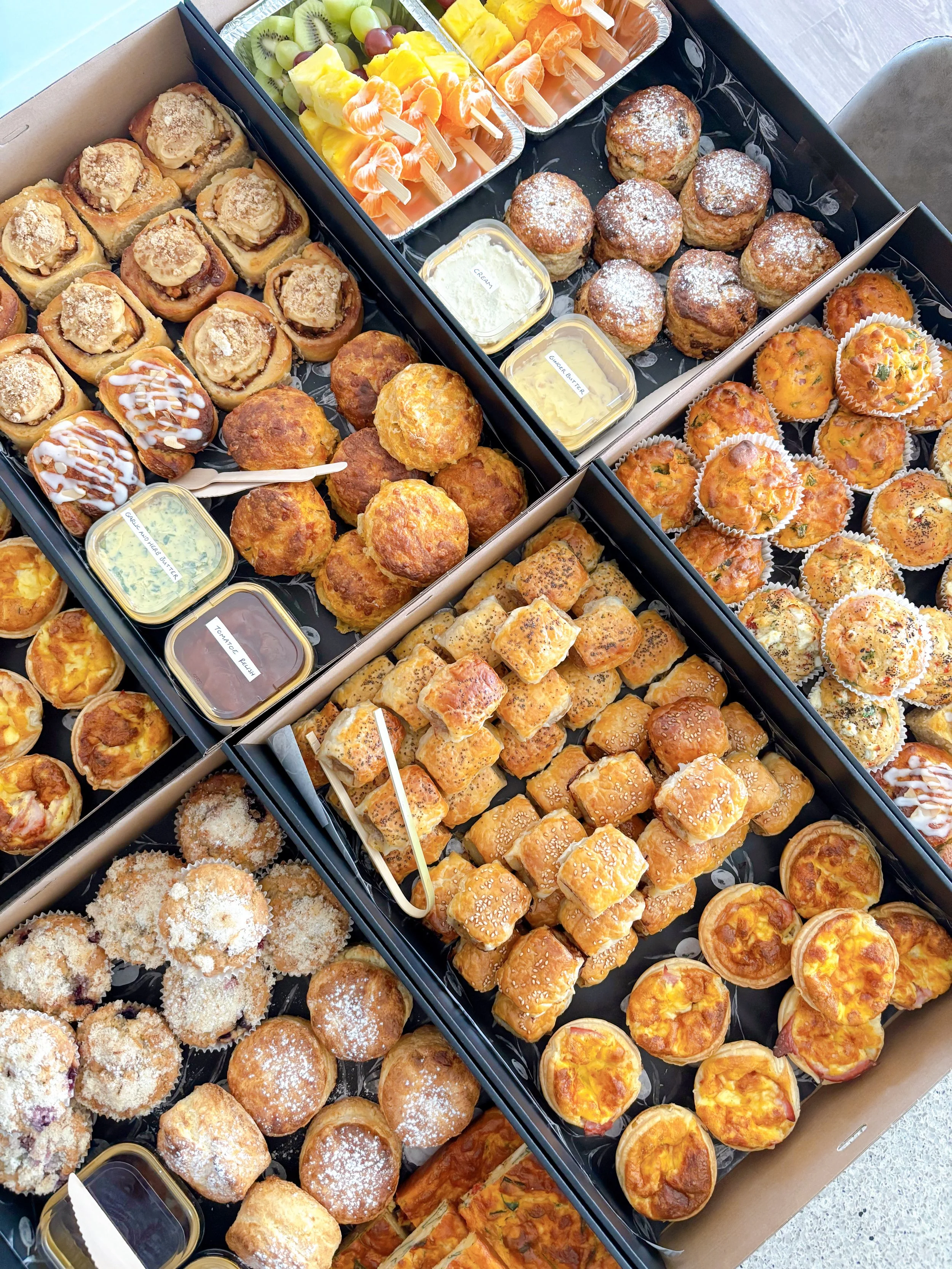 Assorted baked goods and snacks arranged in black trays, including mini quiches, muffins, and finger foods, with small containers of dipping sauces, pulled together in a large cardboard box.