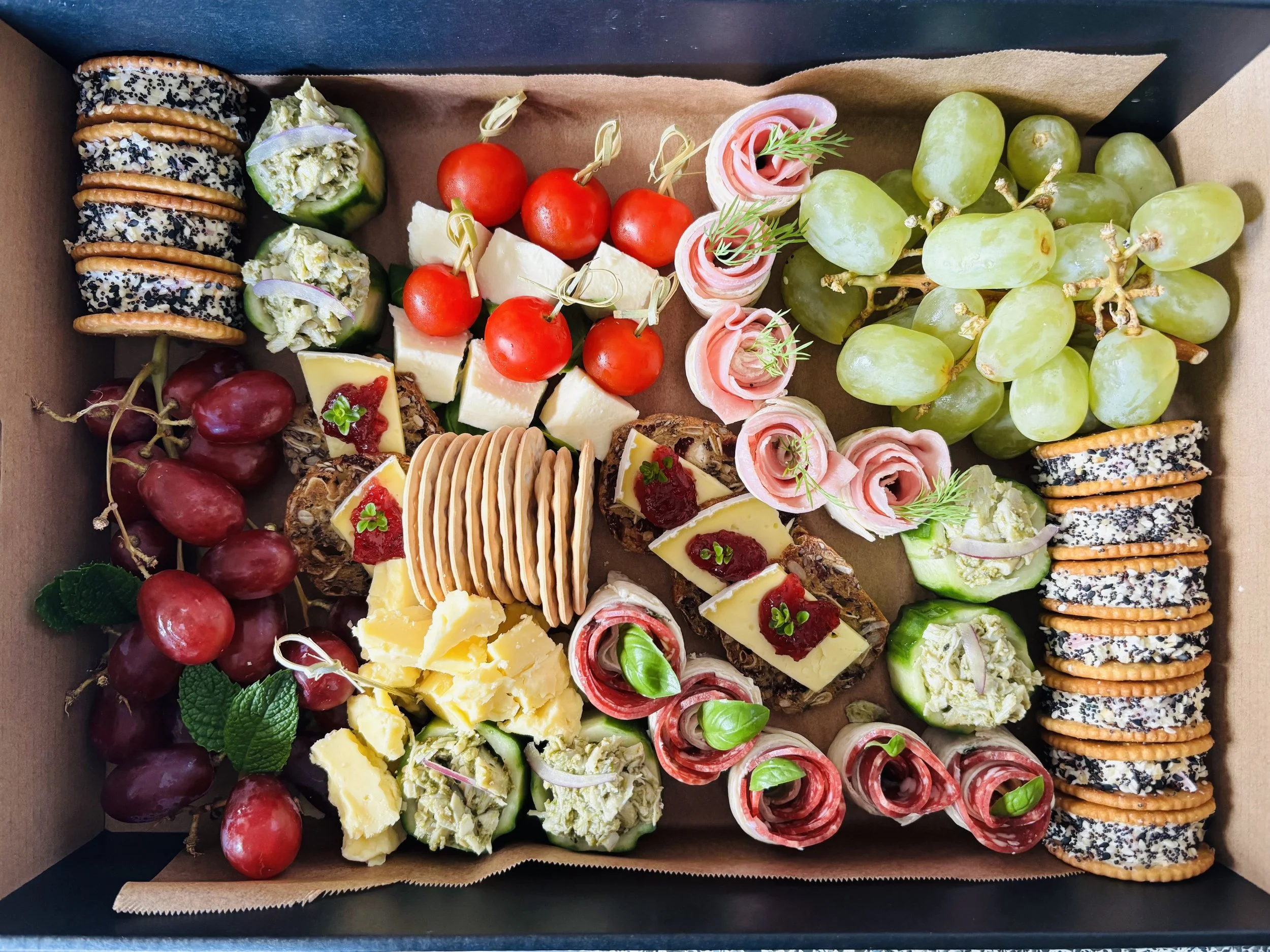 Assorted fruit platter with cherry tomatoes, grapes, cheese, crackers, cucumber bites, and rolled prosciutto with herbs arranged in a box.