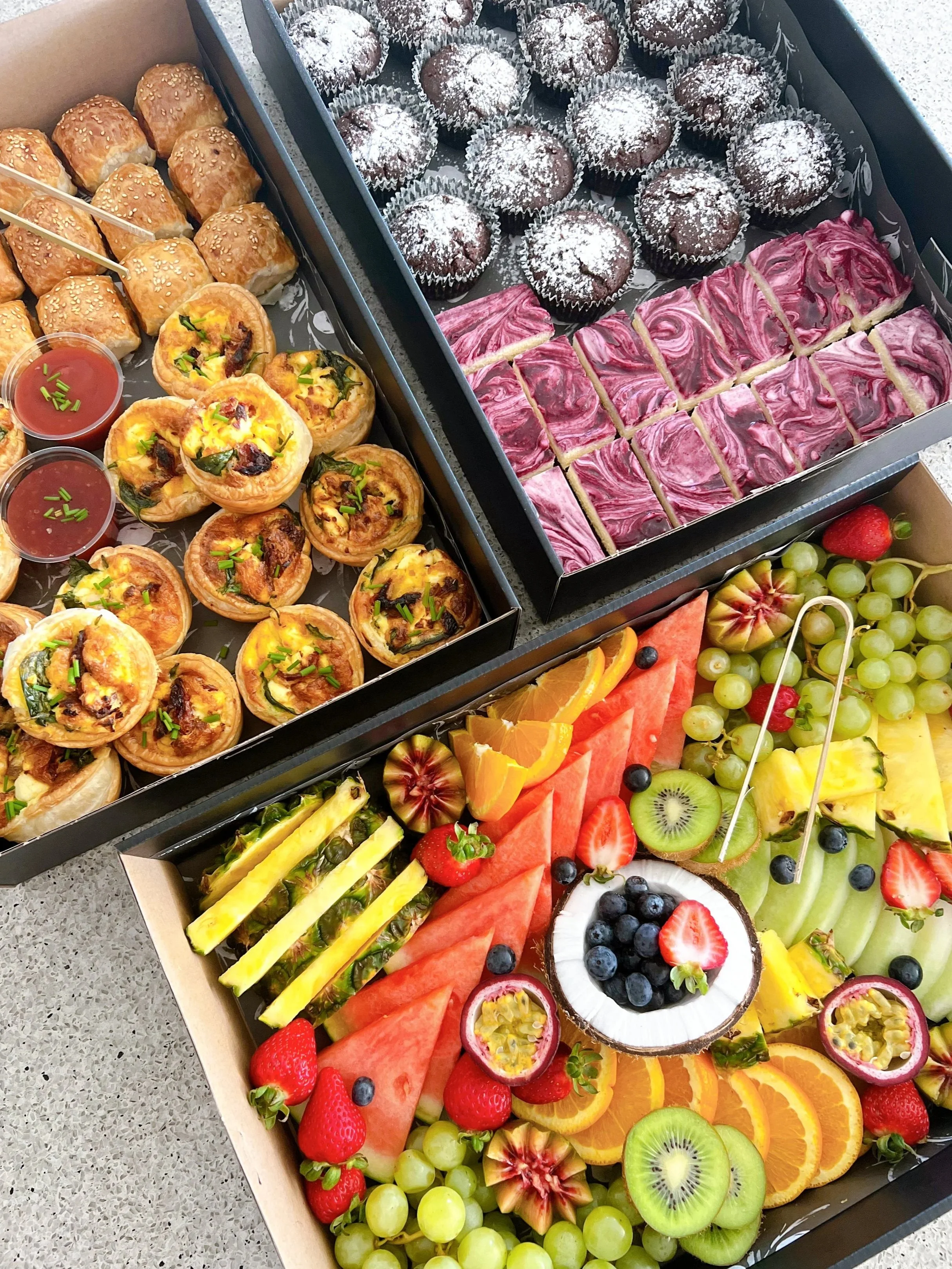 Assorted desserts and fresh fruit arranged in boxes, including mini cheesecakes, chocolate muffins, fruit slices with strawberries, kiwi, pineapple, watermelon, grapes, oranges, and passion fruit.