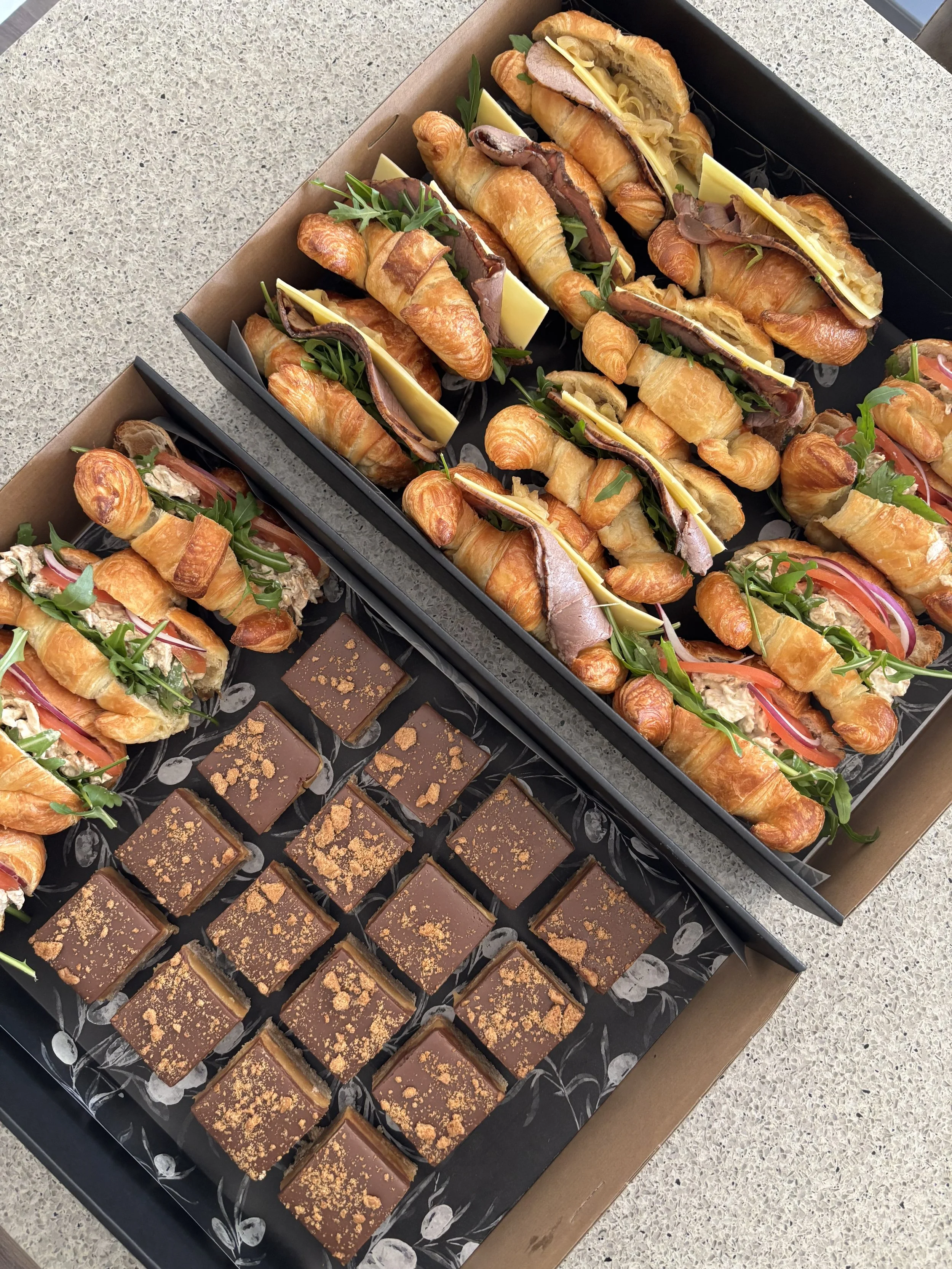 Assorted croissant sandwiches with meats, cheese, and greens, and trays of chocolate squares with caramel topping.