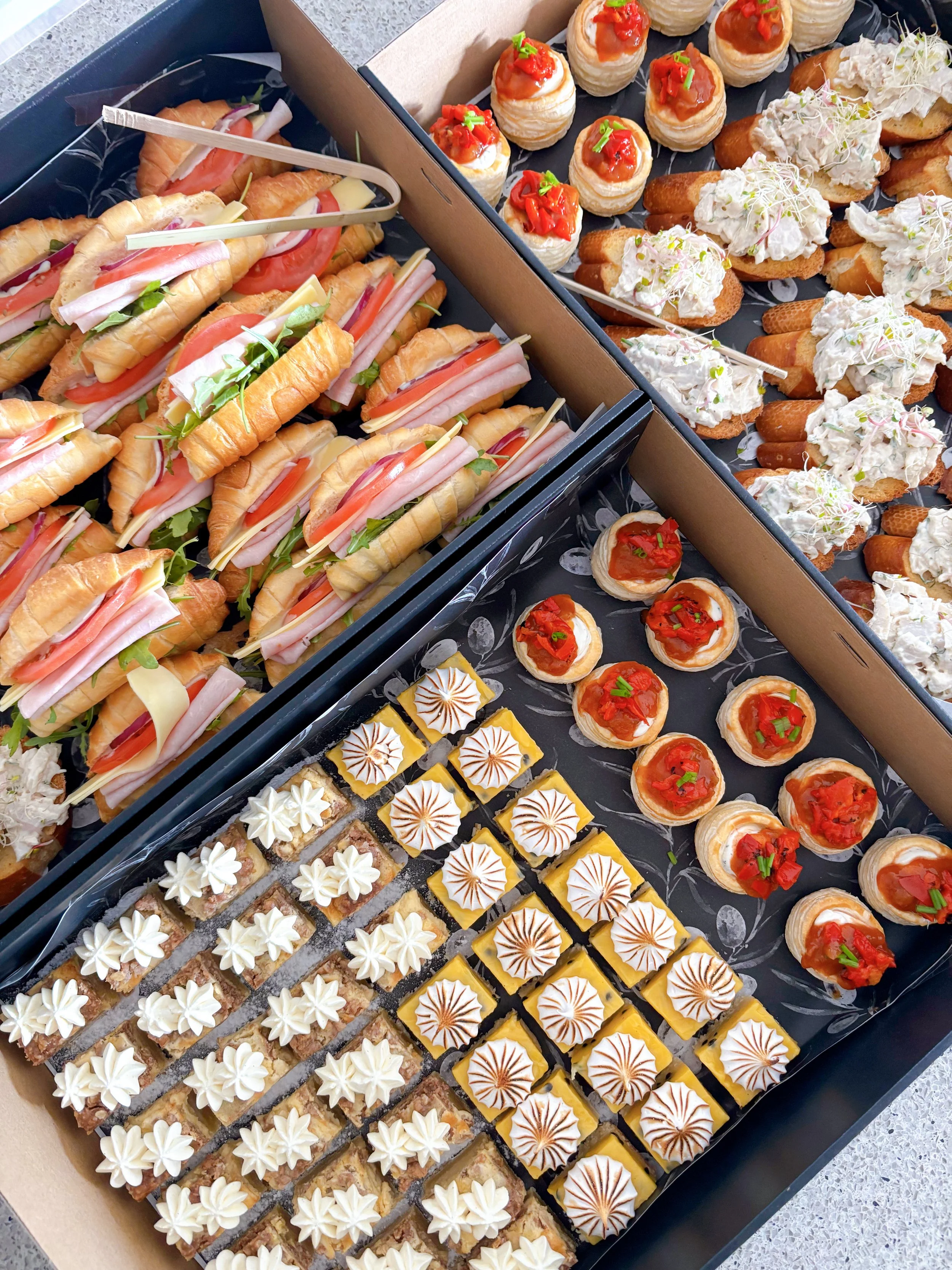 Assorted appetizer tray with mini sandwiches, crostinis topped with creamy spread and red topping, and small desserts with white and gold frosting.