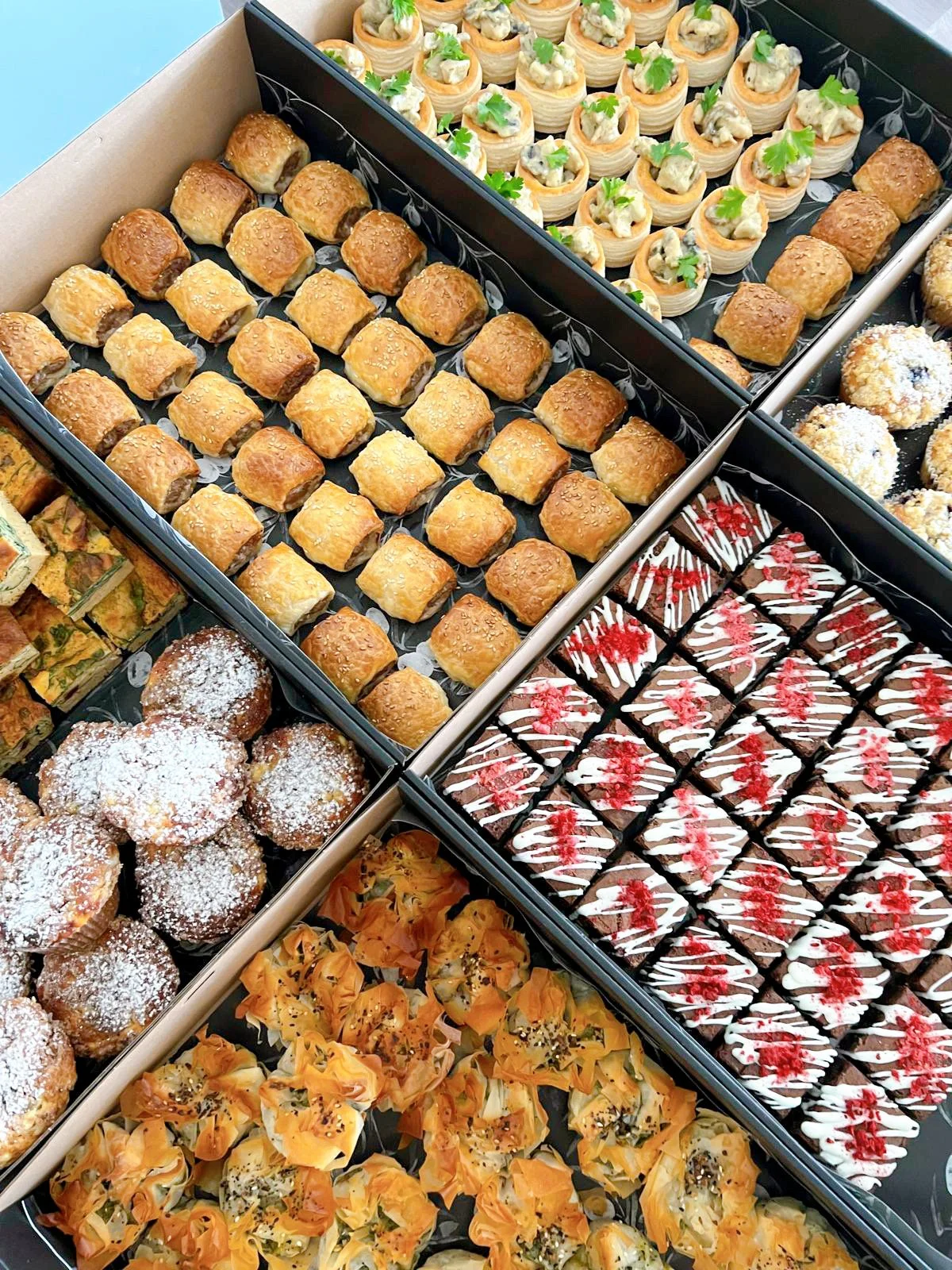 Assorted bite-sized desserts in black display trays, including mini sandwiches, powdered sugar muffins, fruit tartlets, chocolate and white drizzled brownies, and baked pastry cups topped with herbs and seeds.