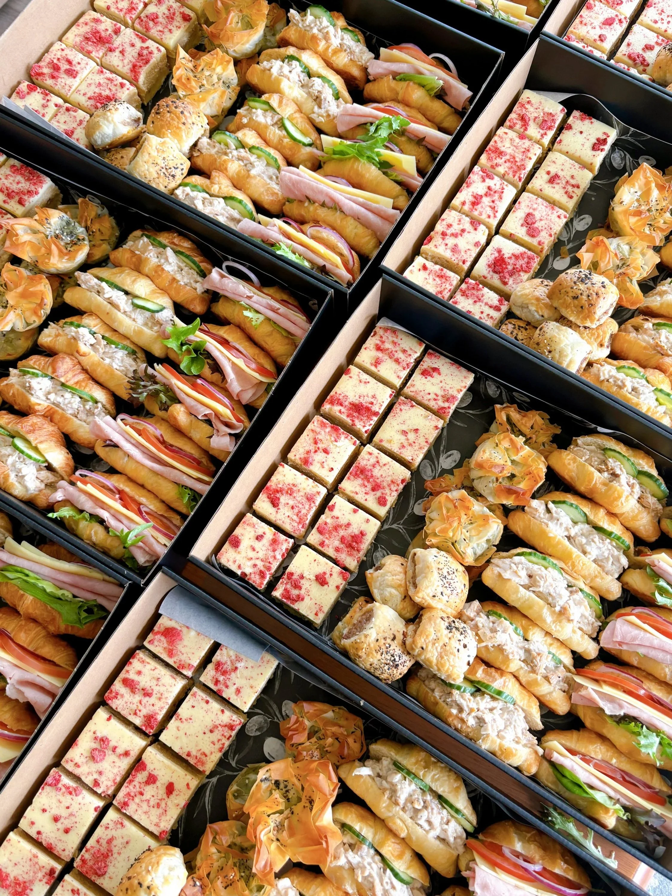 Assorted sandwiches and dessert bars in black boxes, including croissant sandwiches with deli meats and vegetables, and rectangular pink and white decorated cake pieces.