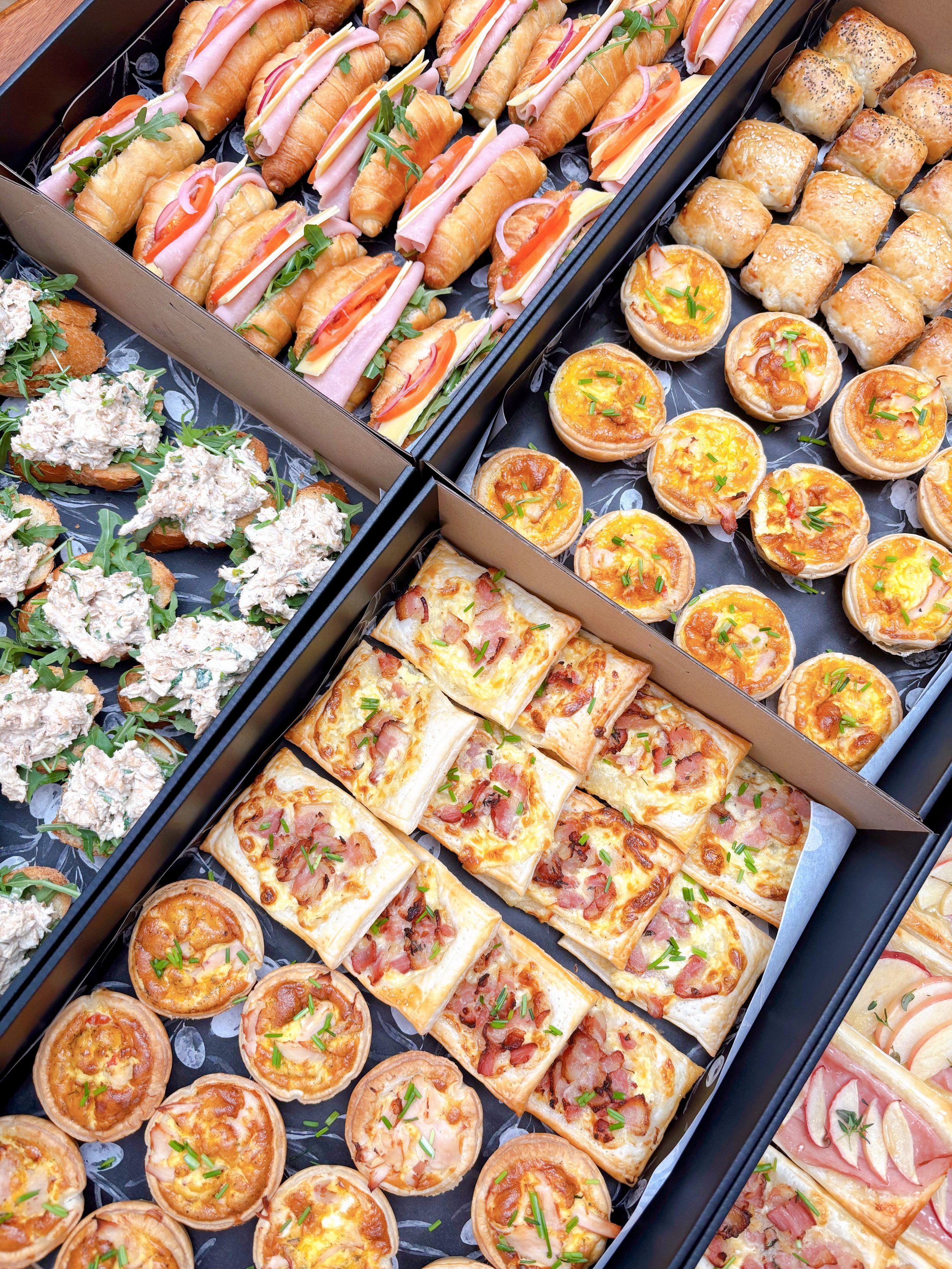 A variety of bite-sized appetizers including croissant sandwiches with ham and cheese, mini quiches, chicken salad crackers, and small bacon pastries.