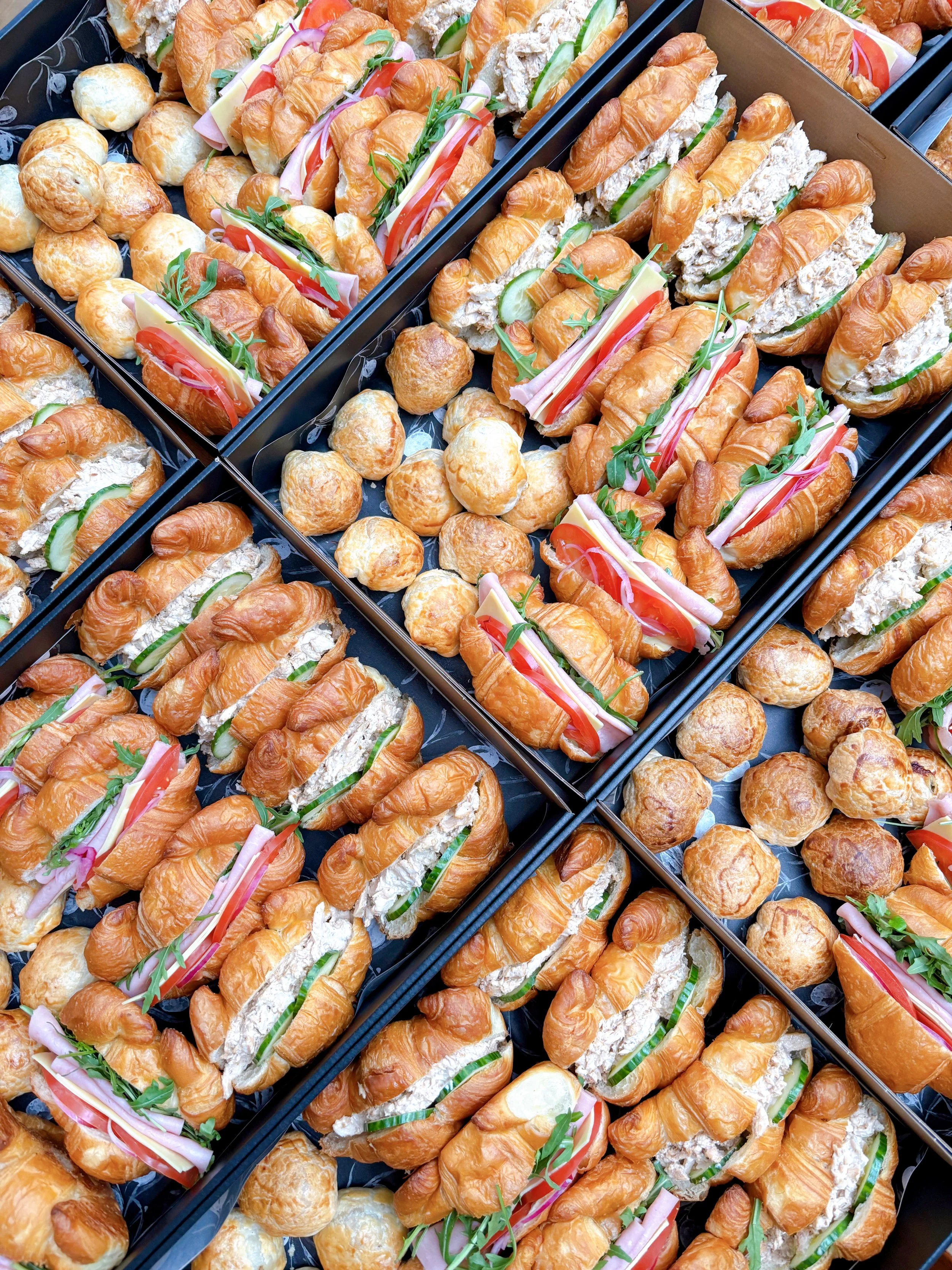 Tray of assorted mini sandwiches and savory snacks including croissant sandwiches with deli meats, cheese, cucumbers, and greens, along with bite-sized cheese puffs.