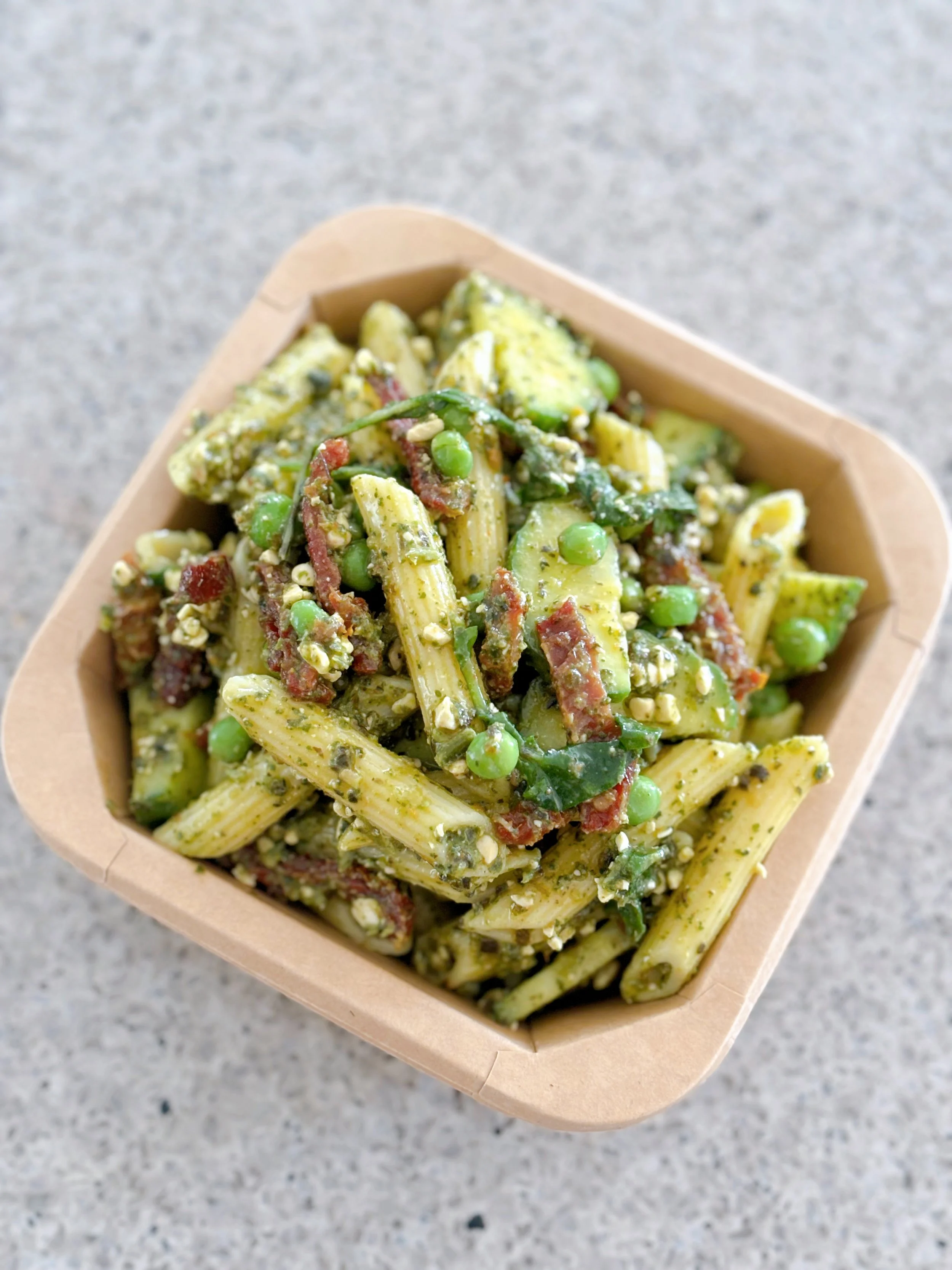 Pasta salad with green peas, chopped bacon, and pesto sauce in a brown container on a gray surface.