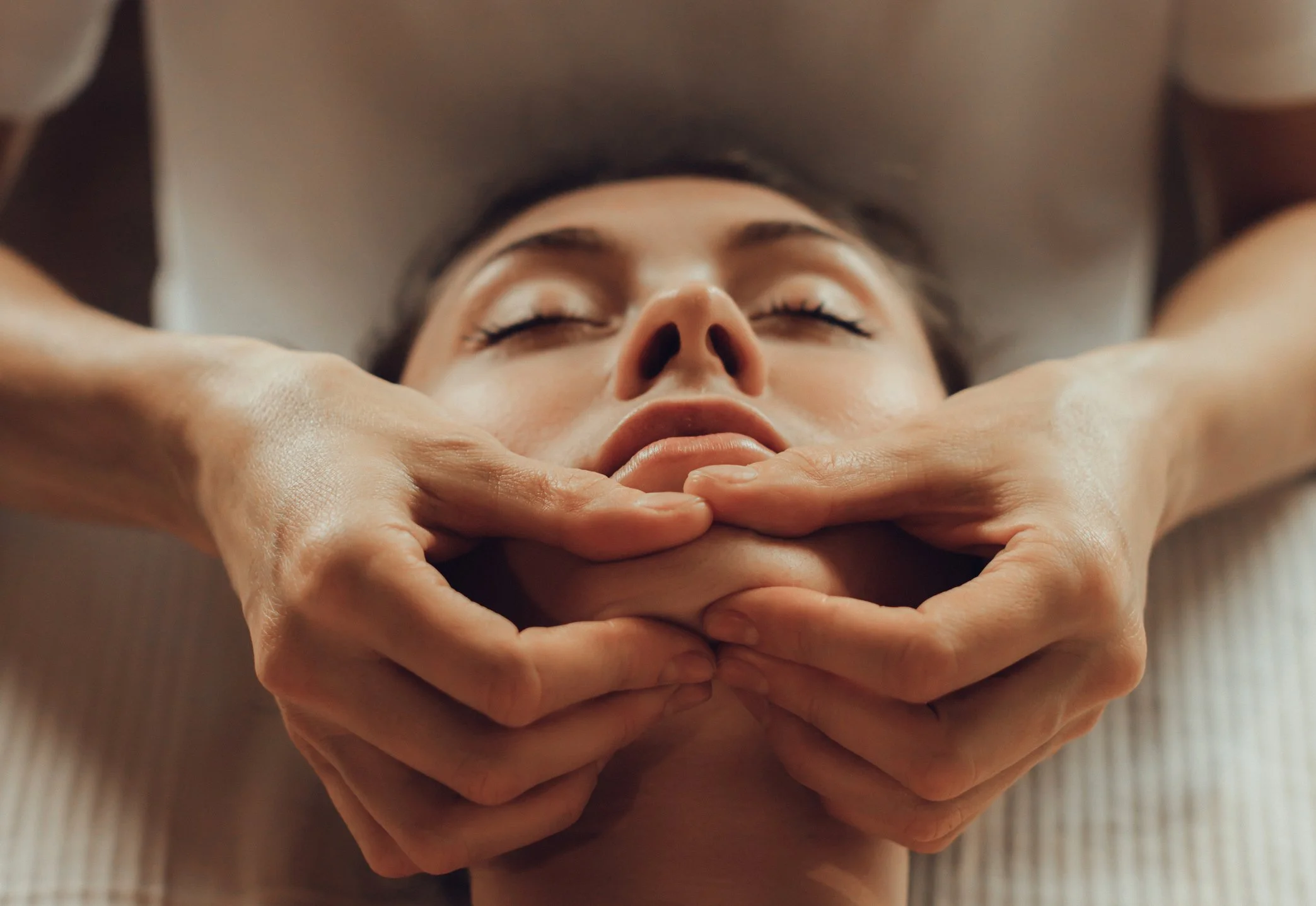 A person with closed eyes receiving a facial massage from a therapist, with hands gently supporting their face and neck.