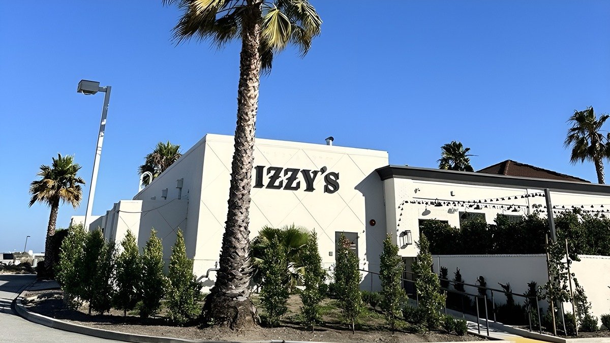 A white building with the sign 'Izzy's' on the front, surrounded by palm trees and bushes, with a clear blue sky in the background.