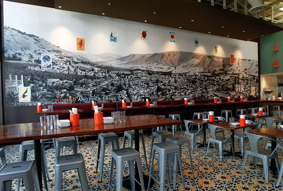 Interior of a restaurant with wooden tables, gray metal stools, orange napkin holders, and a large mural of a cityscape with mountains in the background, and climate-themed artwork on the wall.