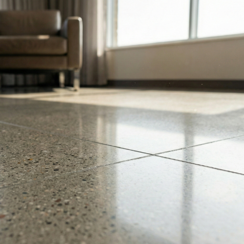 Close-up of a polished tile floor with sunlight coming through a large window, part of a living room with a brown sofa.