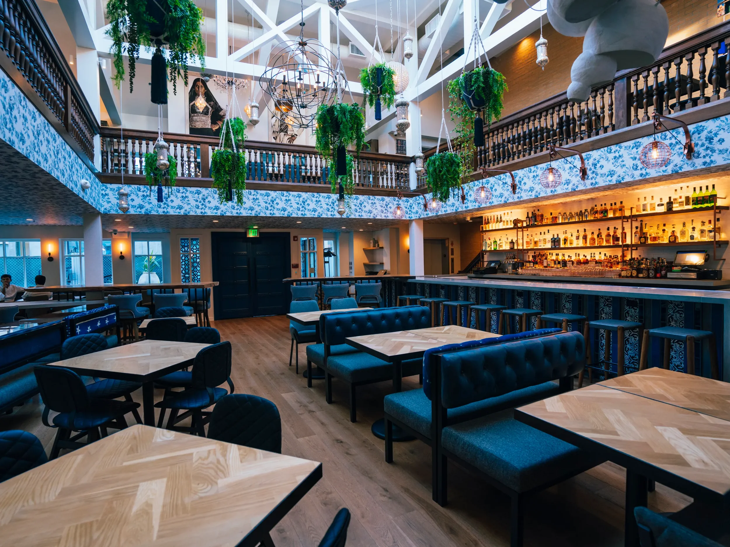 Interior of a stylish bar and restaurant with wooden floors, black and blue furniture, hanging plants, and an illuminated liquor display behind the bar. A second-floor balcony with hanging plants overlooks the space, with the ceiling featuring large spherical light fixtures and artwork on the walls.