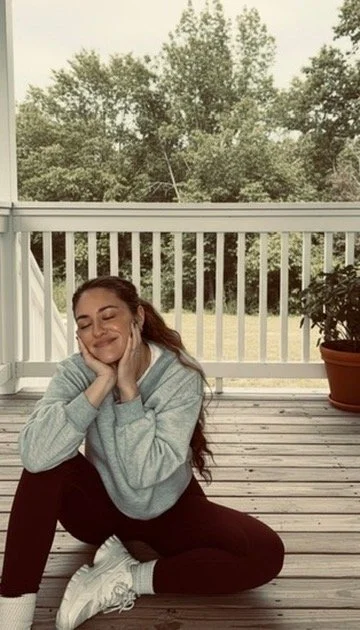 Woman sitting on a wooden porch, smiling with eyes closed, wearing a gray hoodie, white sneakers, and black pants, with green trees in the background.