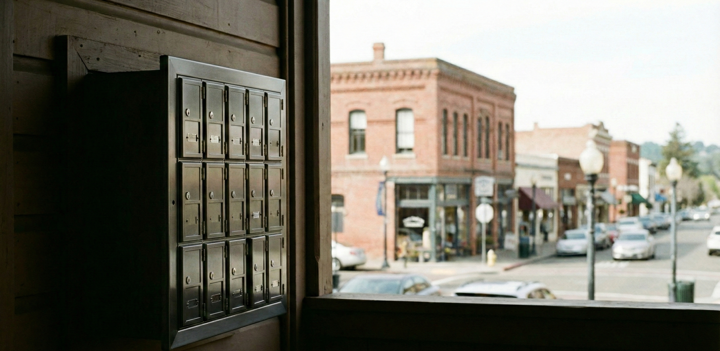 A wall of metal mailboxes inside a building, with a street view through a window showing historic brick buildings, parked cars, street lamps, and sidewalk storefronts in a small town.