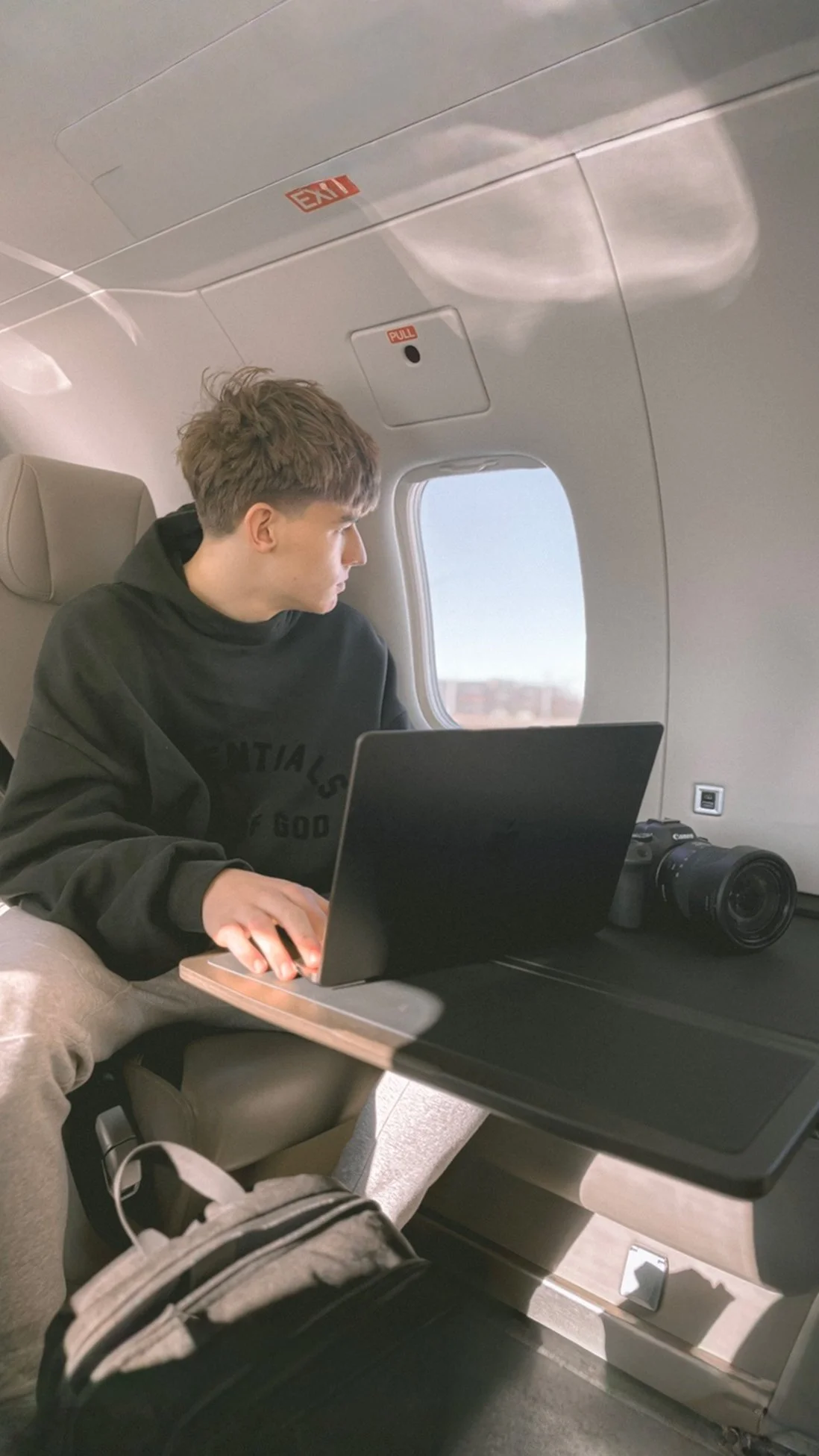 A young man sitting on an airplane seat, working on a laptop placed on a tray table. A camera is beside him. The airplane window shows a blurry landscape outside.