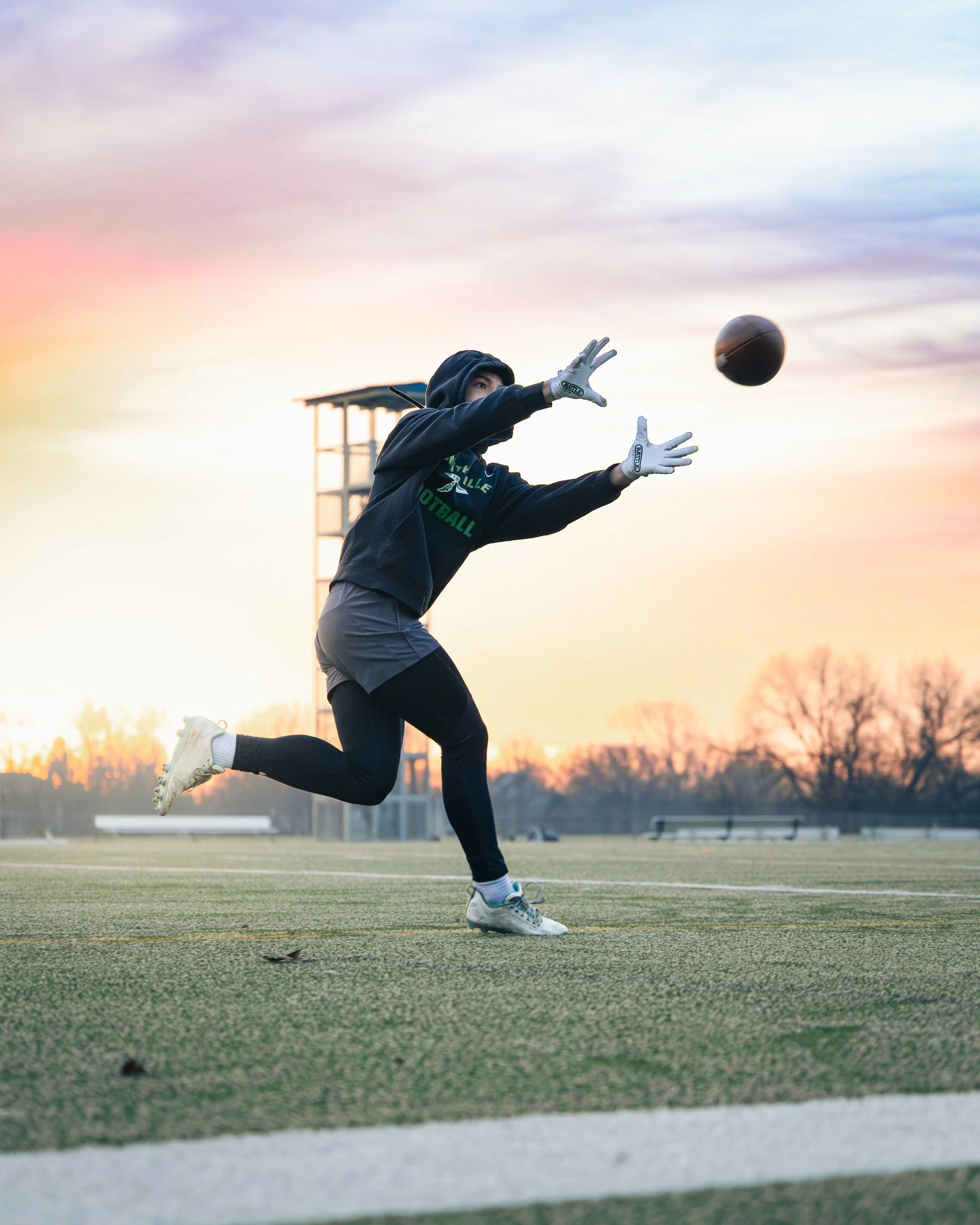 A football goalkeeper in a black hoodie and gray shorts reaching out to catch a football during sunset on a sports field.