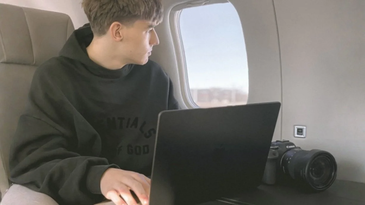 A young man sitting in an airplane seat, working on a laptop, with a camera placed beside him.