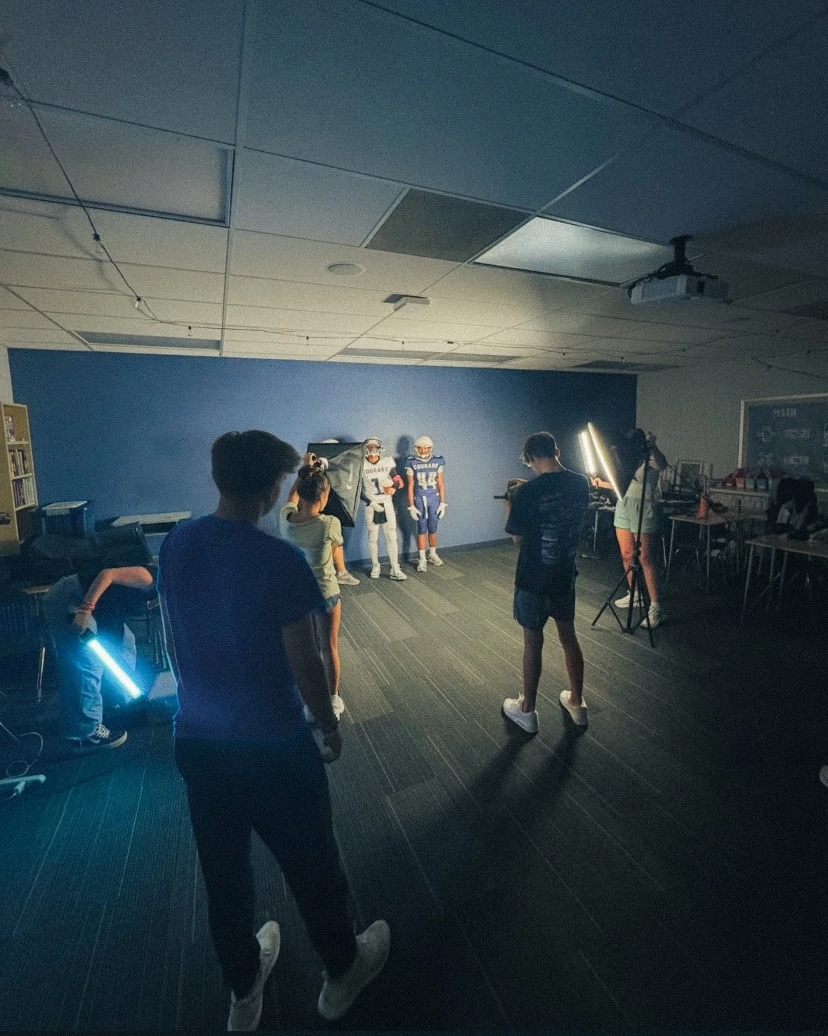 A group of people is filming a sports scene with two football players in a classroom or studio setting, while others hold lights and equipment.