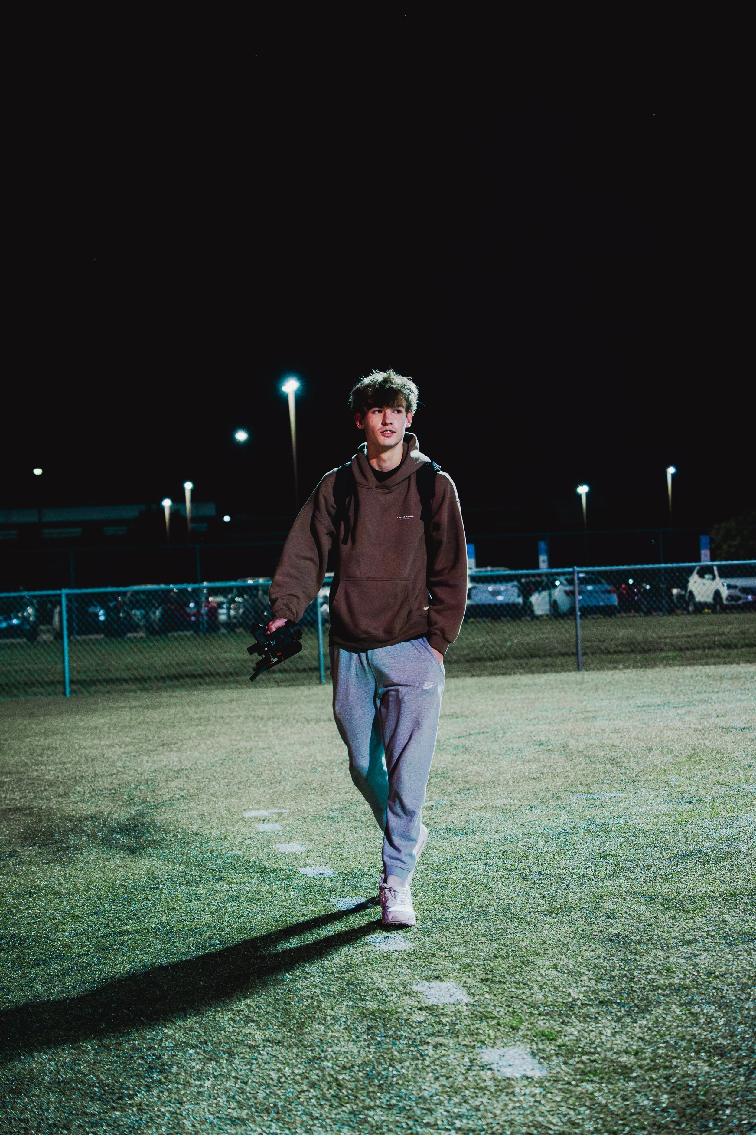 A young man dressed in a brown hoodie, gray sweatpants, and white sneakers walking on a sports field at night, holding a camera, with parking lot and streetlights in the background.
