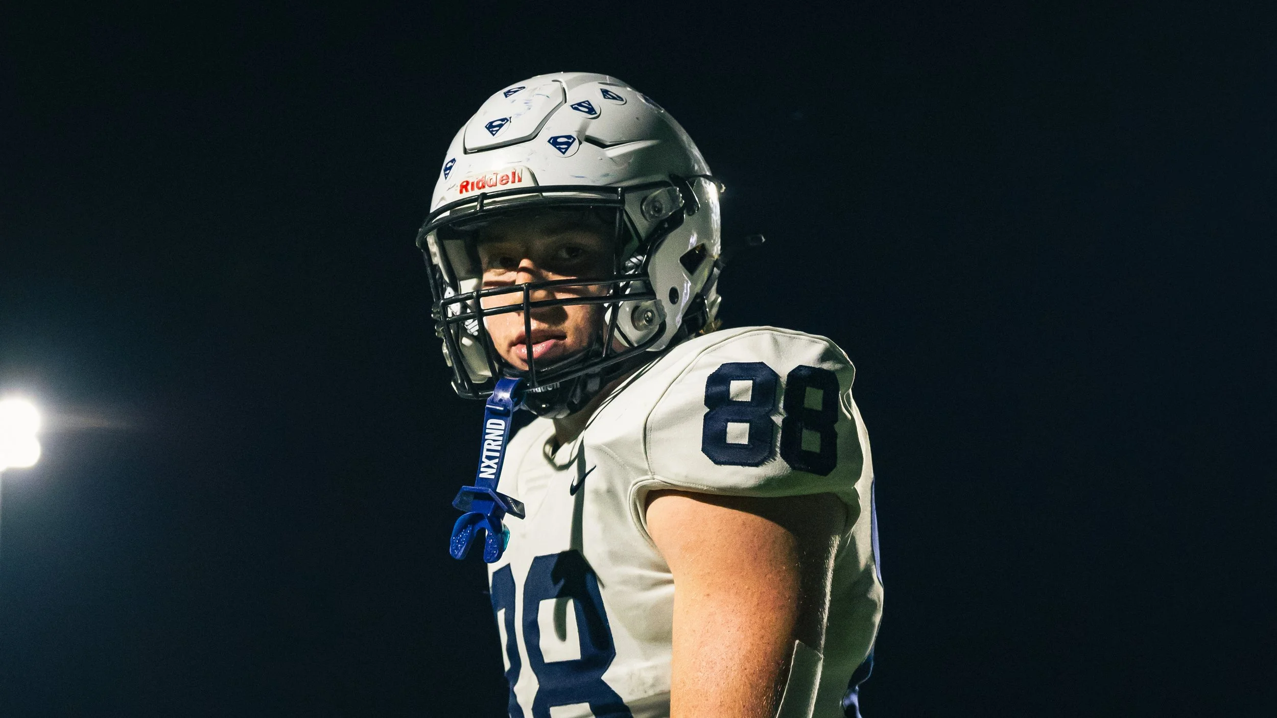 A football player wearing a white helmet with a face mask, a white jersey with navy numbers 88, and a blue mouth guard hanging from his face mask, standing against a dark night sky with stadium lights in the background.