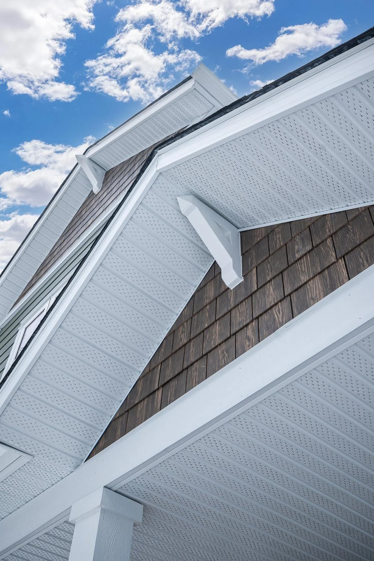 Close-up of a house roof showing white soffit and fascia, brown shingle siding, and a blue sky with clouds.