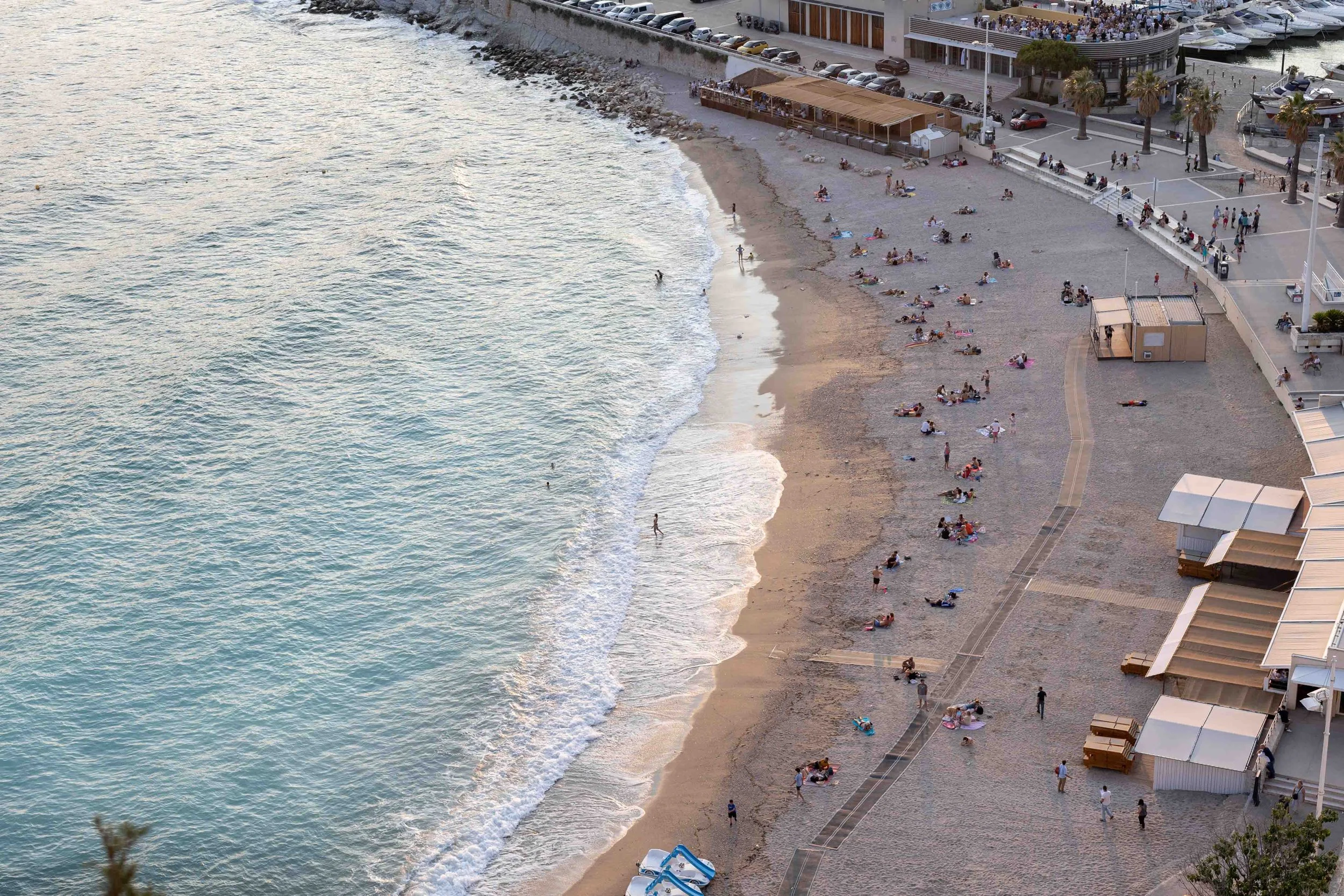Une plage urbaine avec des personnes qui se reposent, nagent et profitent du soleil, bordée par des constructions modernes et des palmiers.