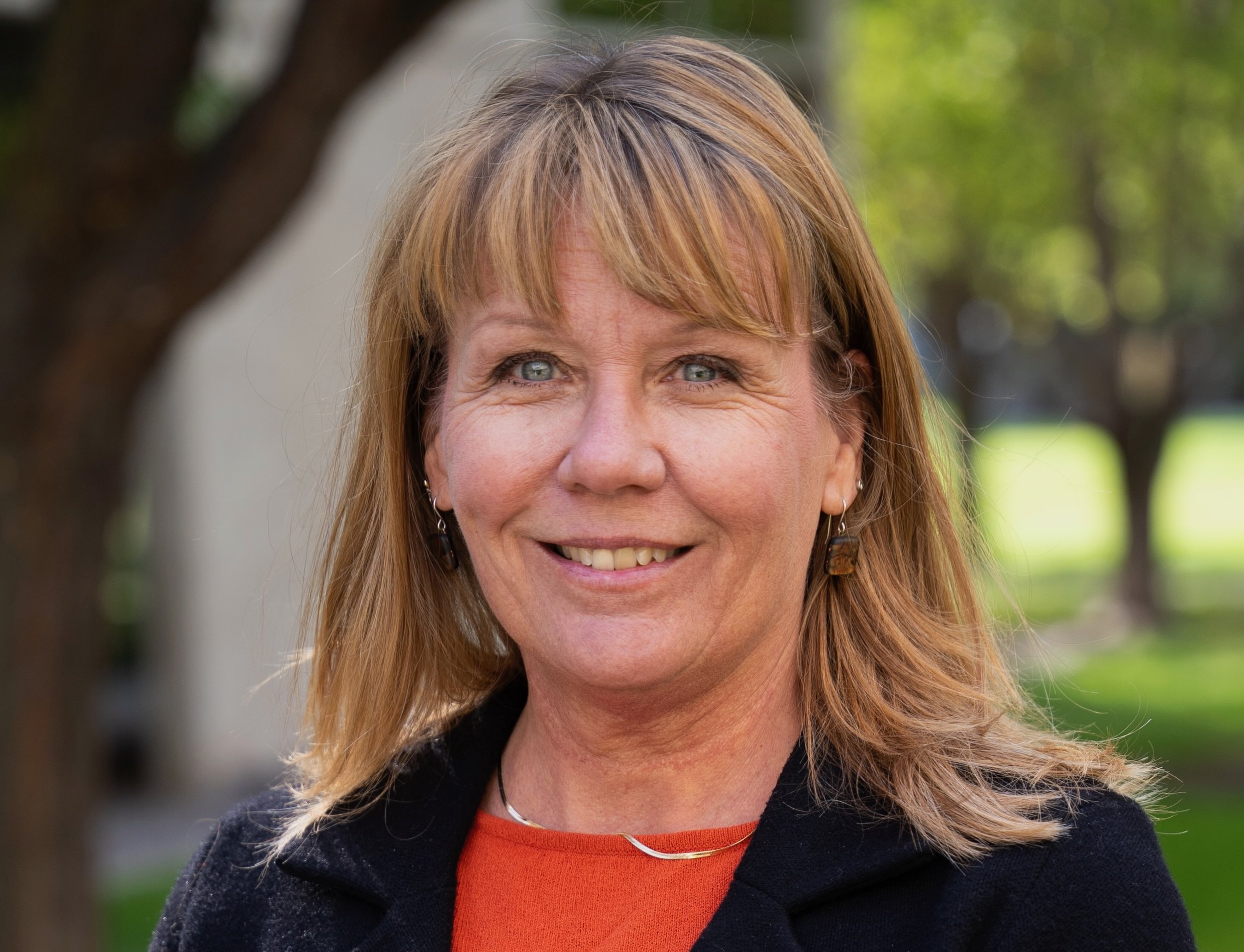 A smiling middle-aged woman with shoulder-length reddish-brown hair and blue eyes, wearing earrings, a black jacket, and an orange top, standing outdoors with trees and greenery in the background.