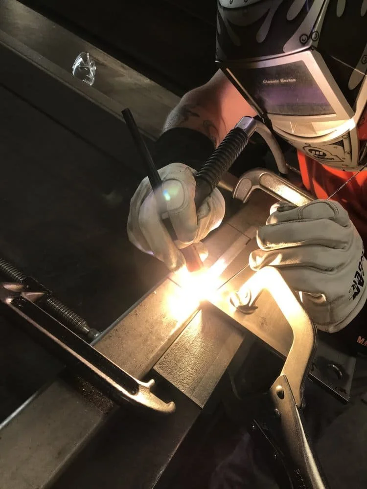 A person welding metal pieces together, wearing a welding helmet and protective gloves, with sparks visible at the welding point.