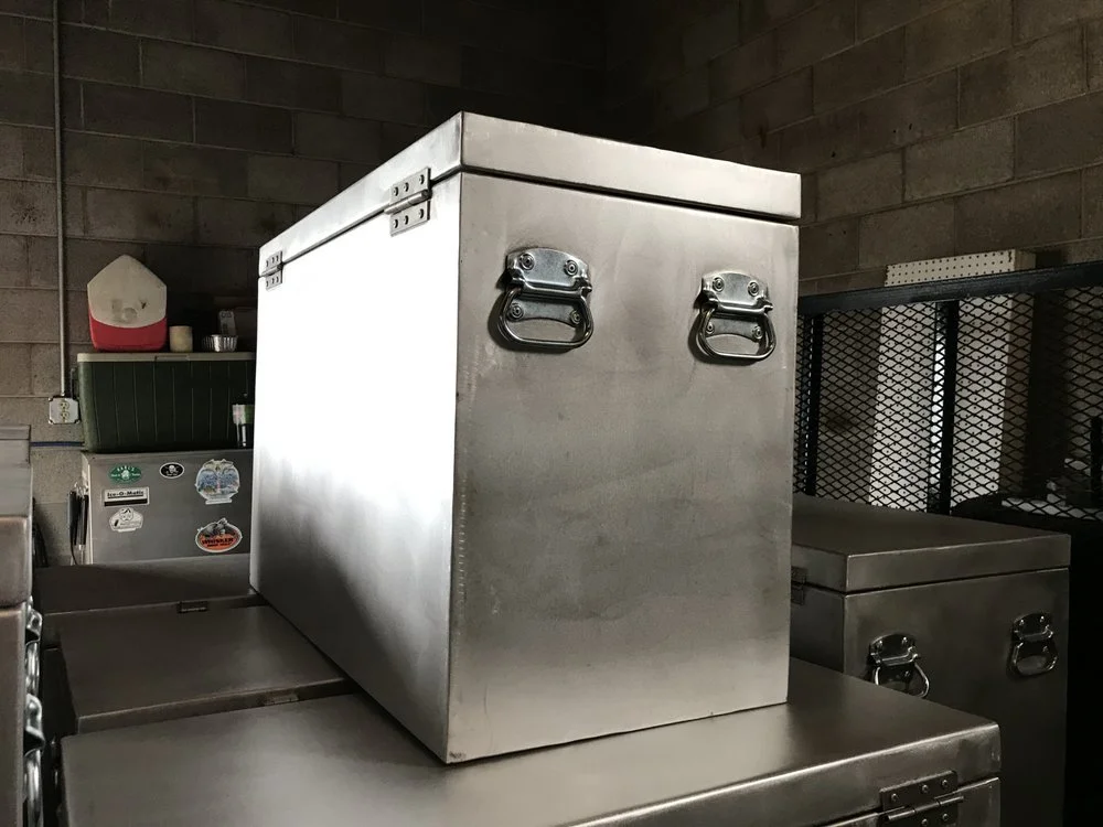 Stainless steel cooler box on a flat surface in a storage room with brick walls and other containers.