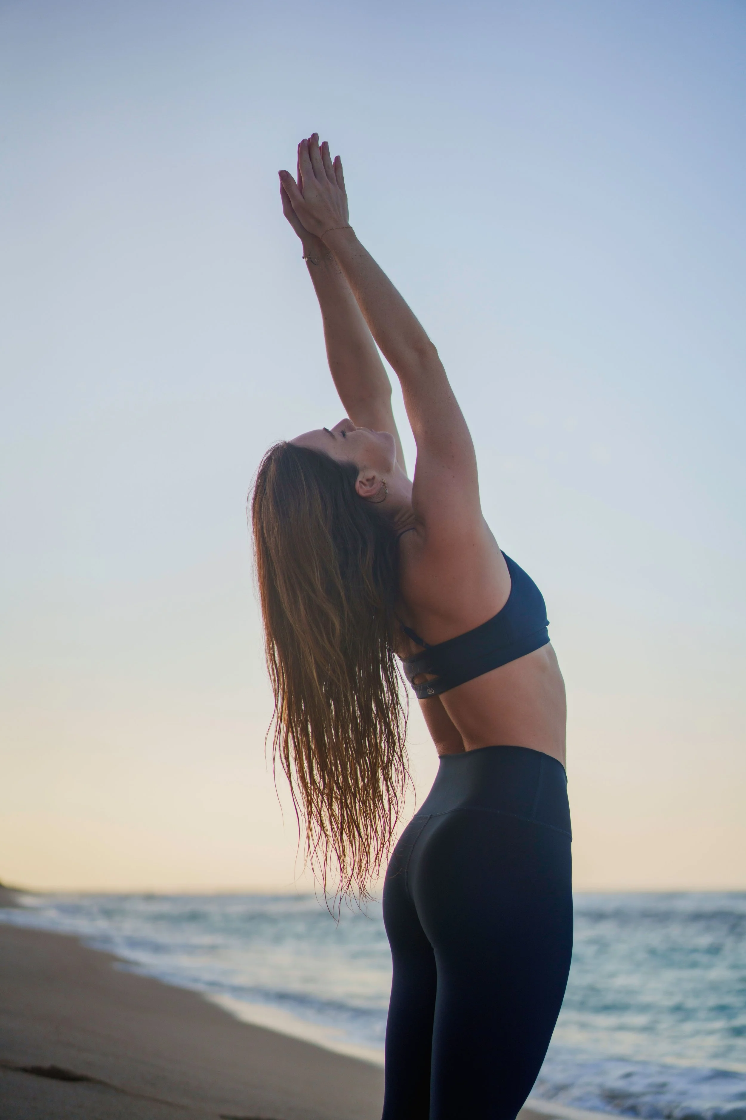 A woman stretching on the beach at sunset, wearing a black sports bra and leggings.