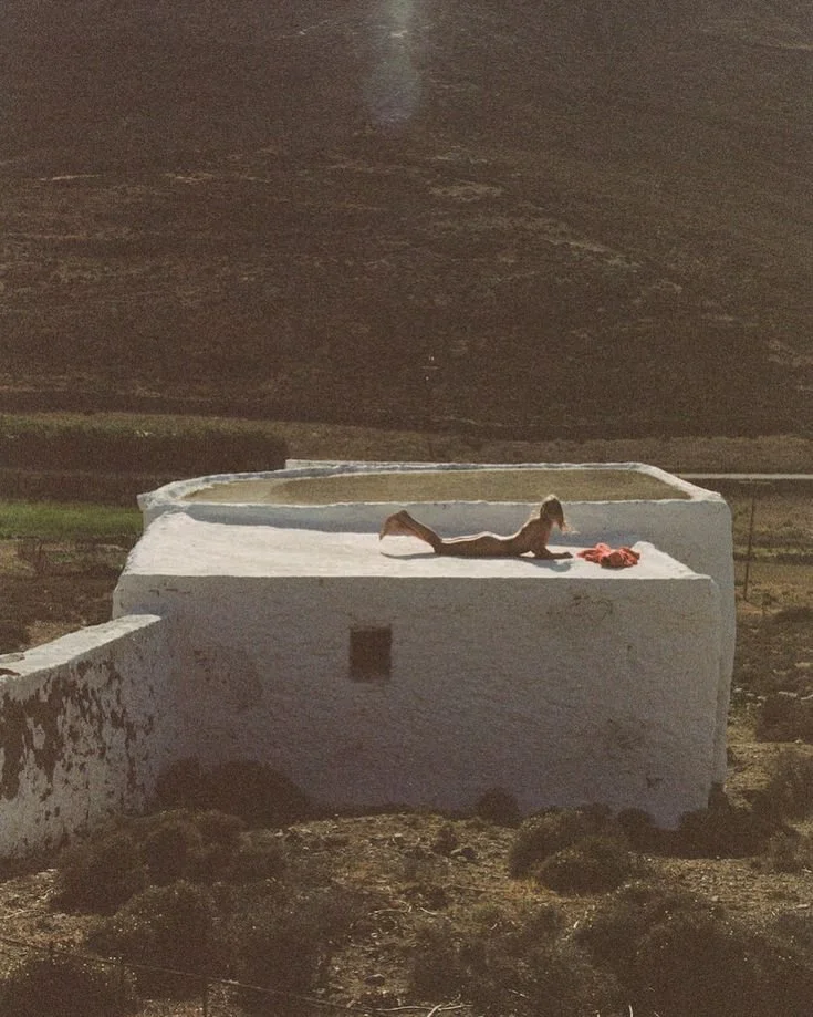 A woman lying on her stomach on the edge of a raised, white, rectangular pool or water container outdoors, with a mountainous landscape in the background.