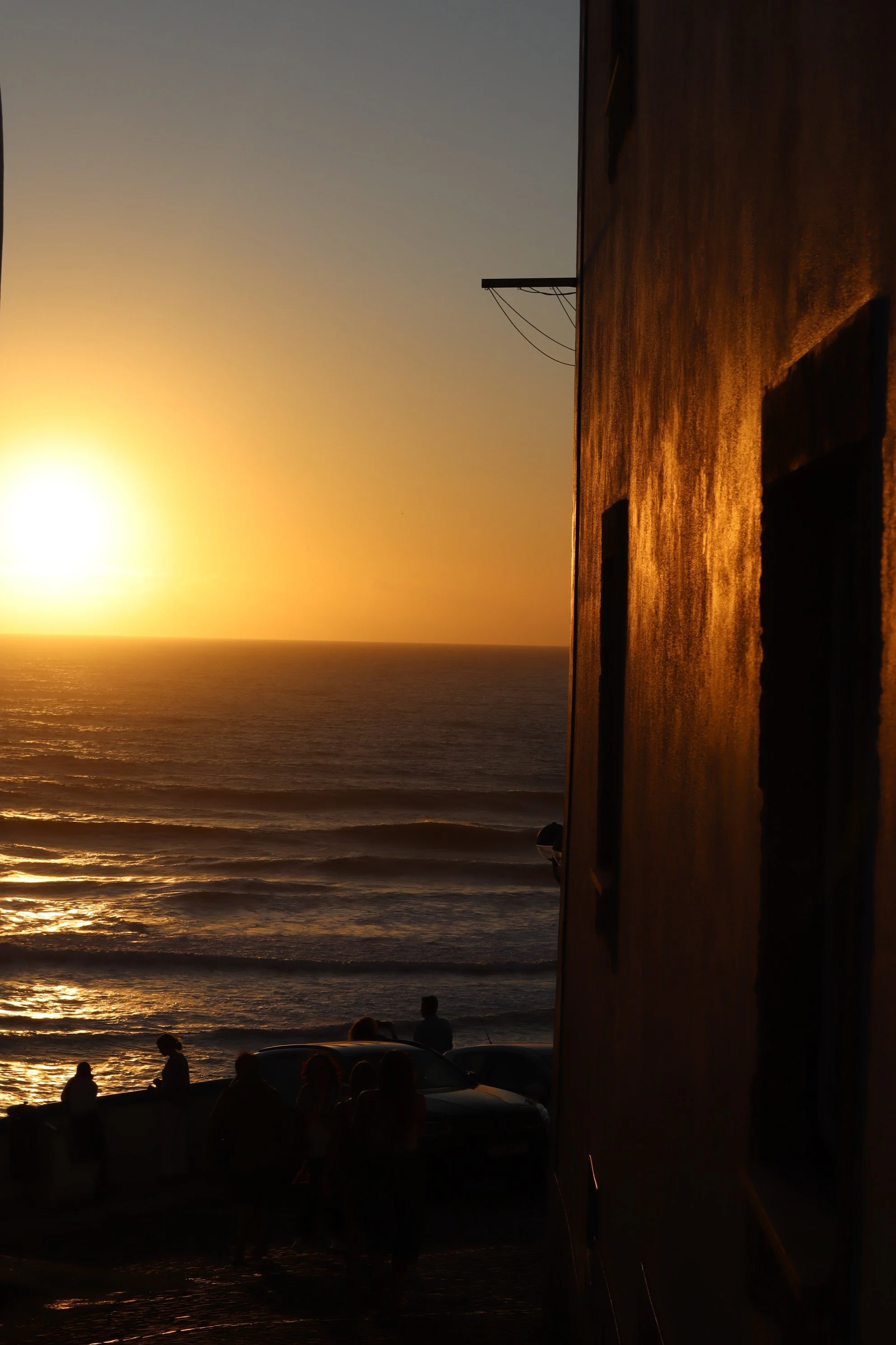 Sunset over the ocean with silhouettes of people and cars near the shoreline, reflected on a building's wall.