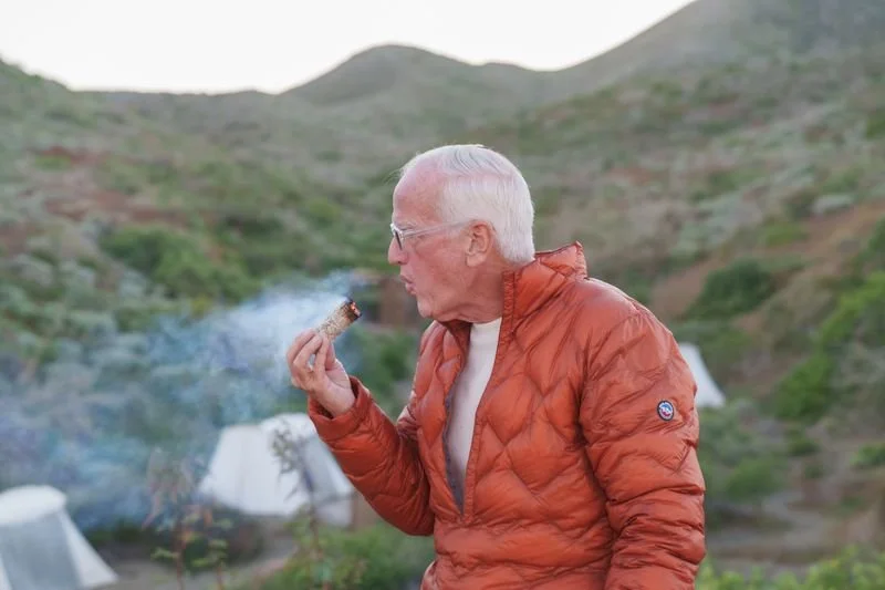 Older man with white hair and glasses, wearing an orange jacket, smoking a cigar outdoors with hills and tents in the background.