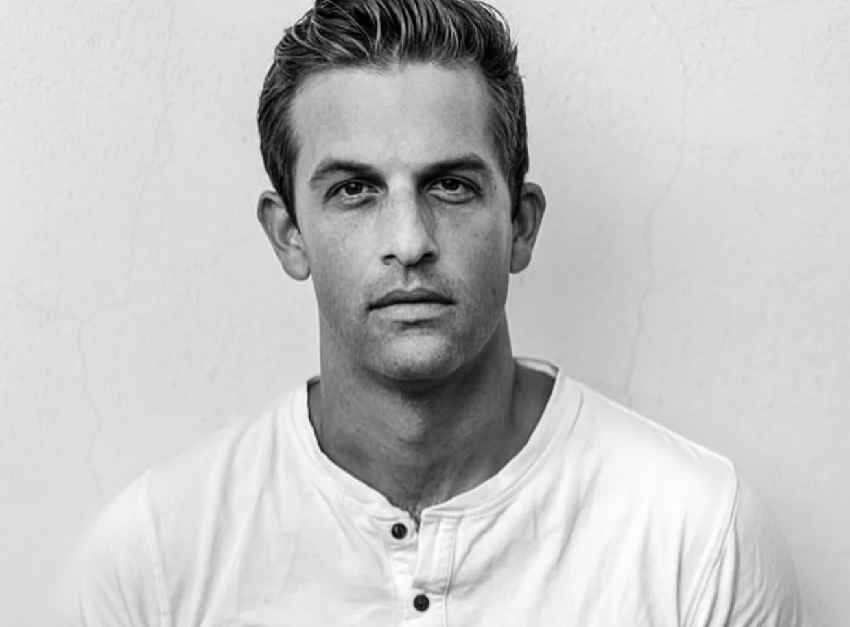Black and white portrait of a young man with short hair, wearing a white shirt, looking directly at the camera in front of a plain wall.