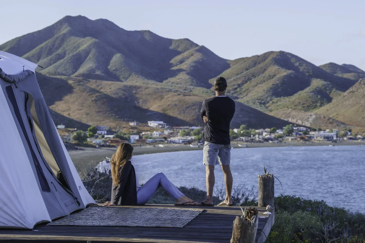 A woman with long hair sitting on a wooden dock next to a tent, and a man standing with arms crossed, looking at a coastal town and mountains in the distance.