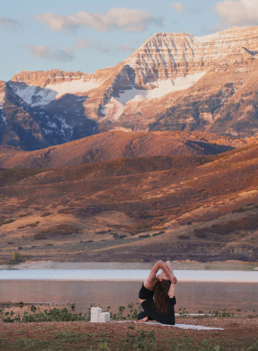 A woman practicing yoga on a mat by a lake with mountains in the background during sunset.