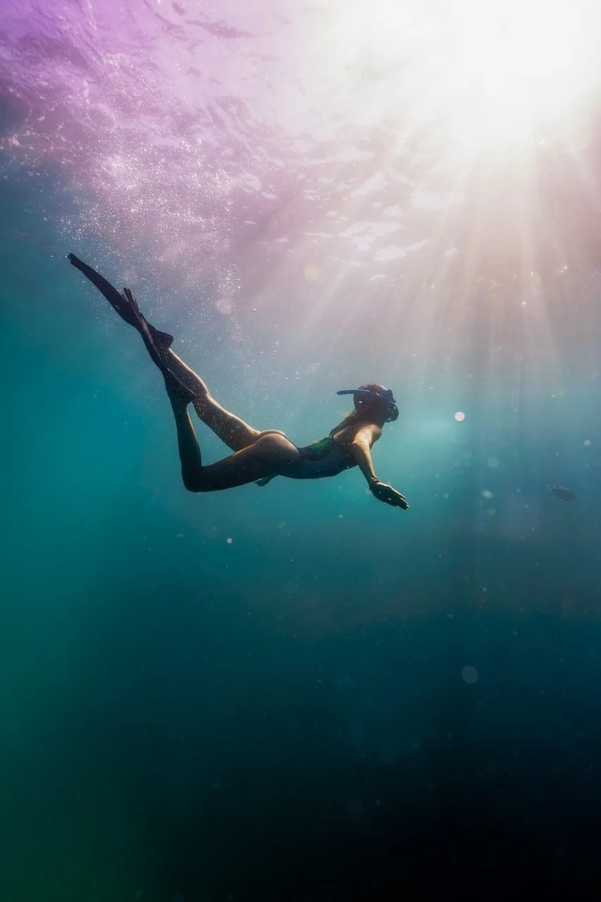 Swimmer in a snorkel mask swimming underwater with sunlight streaming from the surface.