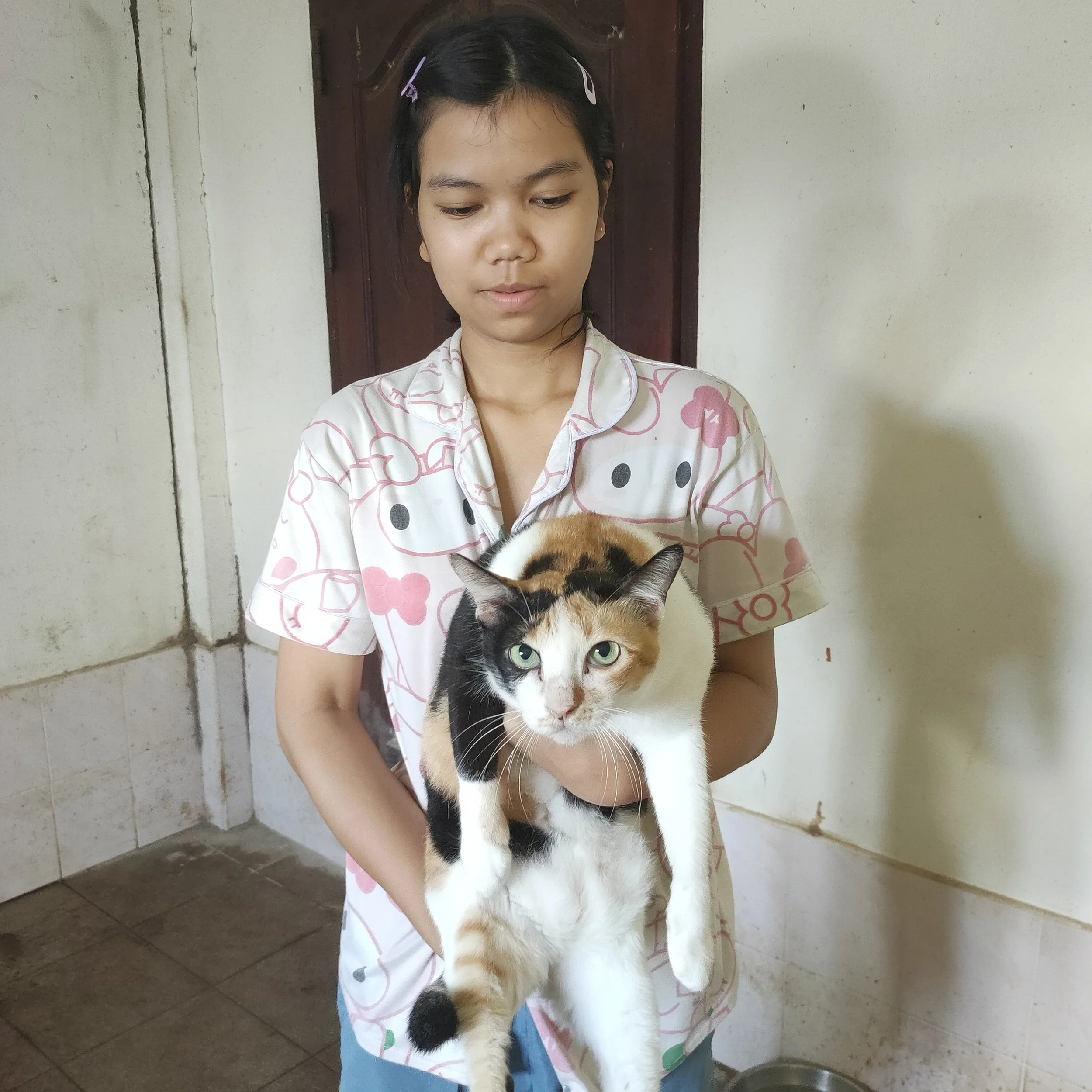 A young girl holding a calico cat indoors against a white wall with a wooden door. The girl is wearing pajamas with a Hello Kitty design and has dark hair clipped back with pink hair clips.