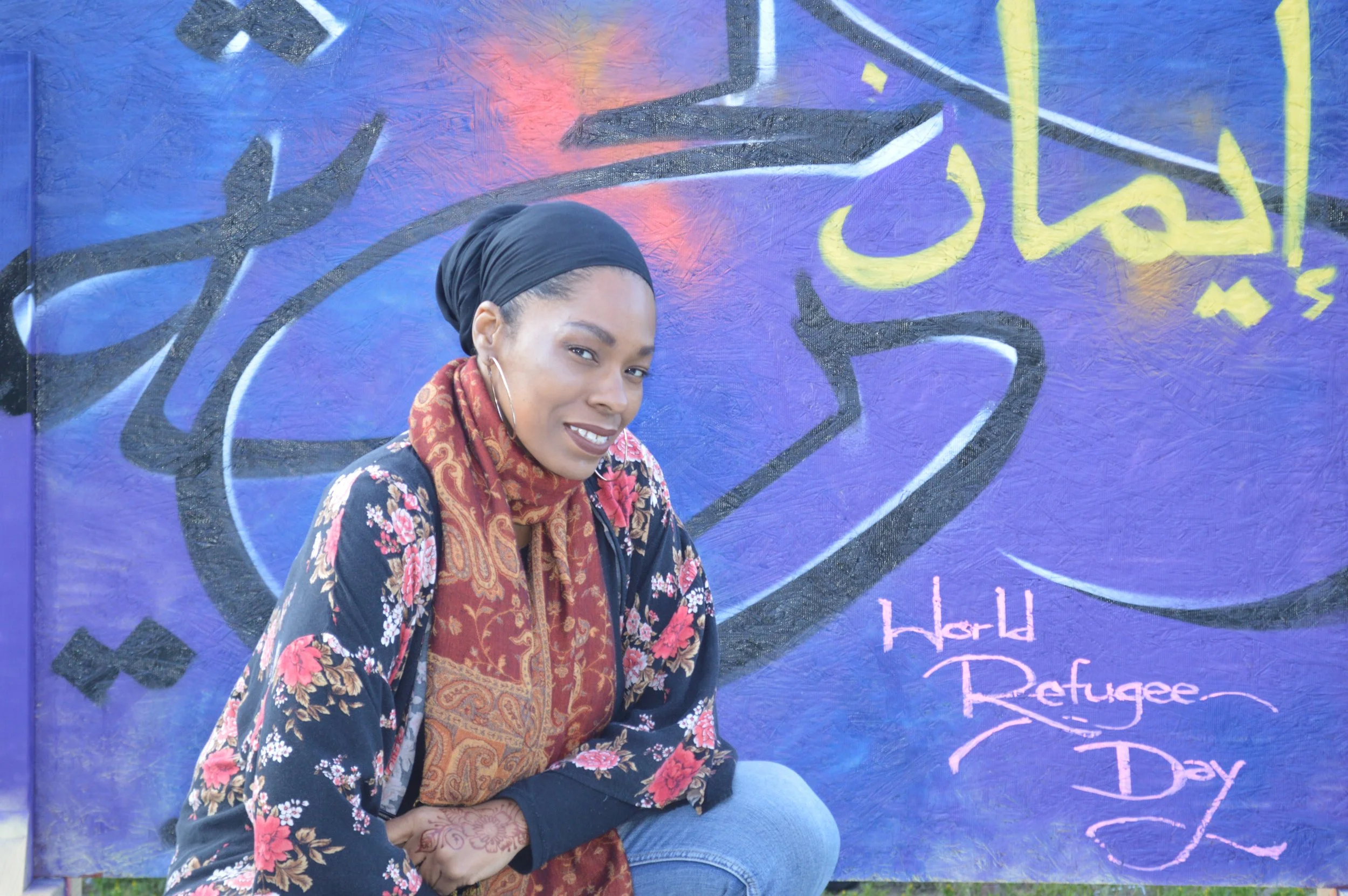A woman sitting in front of a colorful mural that says 'World Refugee Day' in pink, with a background featuring purple, black, yellow, and other colors.