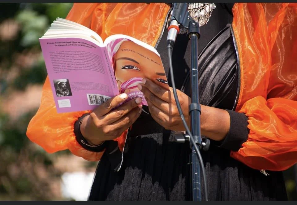 Person reading a pink book with a close-up photo of a woman's face on the cover, outdoors, wearing an orange jacket and black dress.