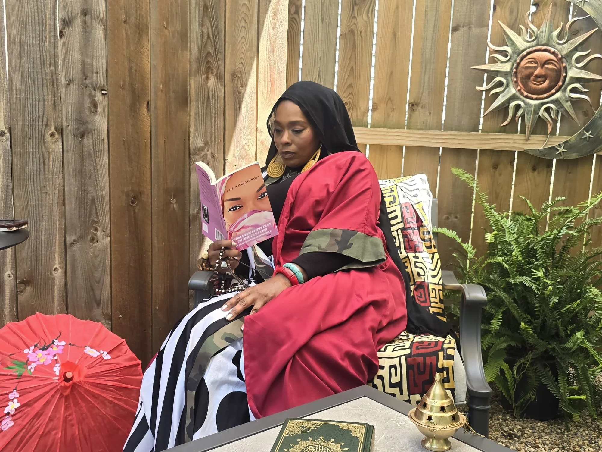 A woman sitting outdoors reading a book, surrounded by decorative items including a red parasol with floral design, a golden ornament, and plants, with a wooden fence and sun and moon wall art in the background.
