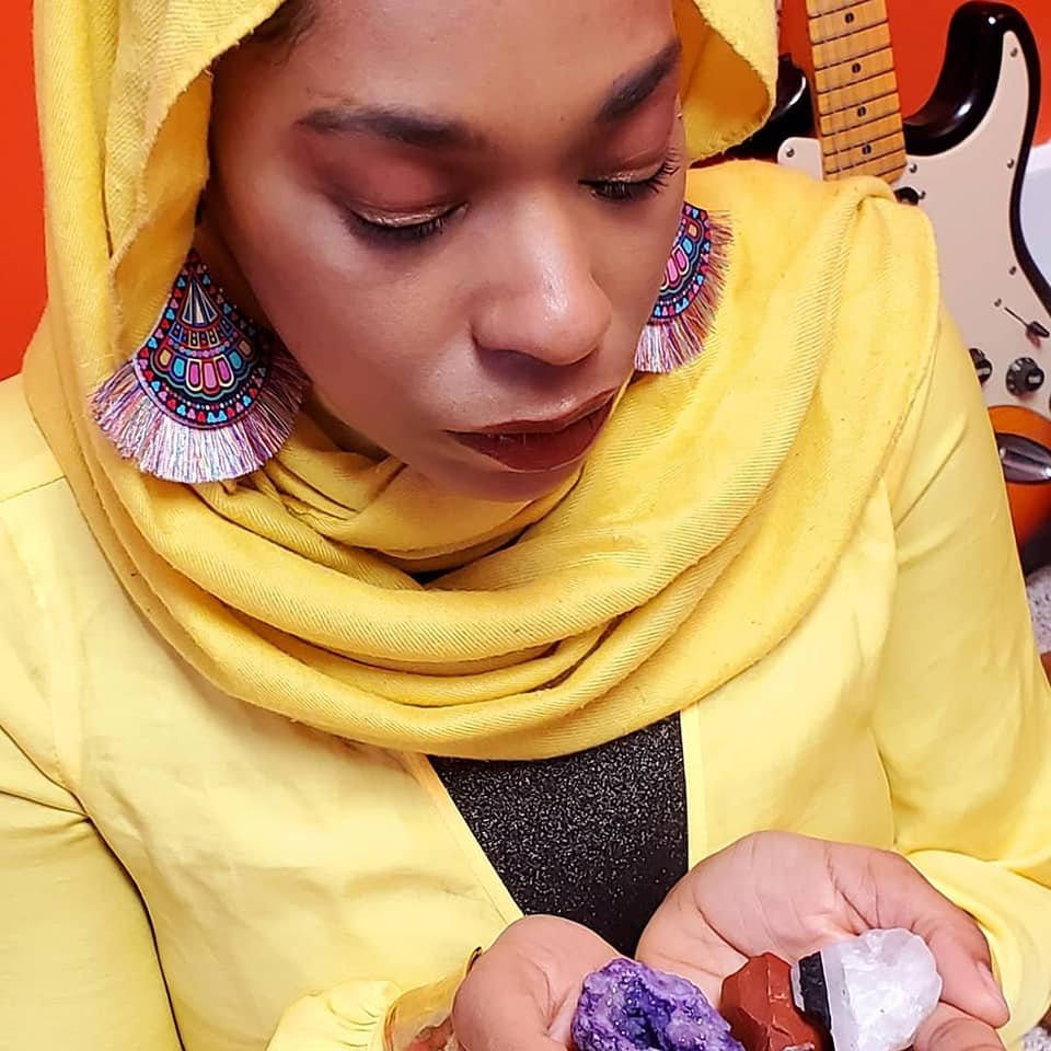 A woman in a yellow hijab and matching yellow top is holding and looking at a small purple and white mineral specimen. She is wearing colorful earrings and is in a room with musical instruments in the background.