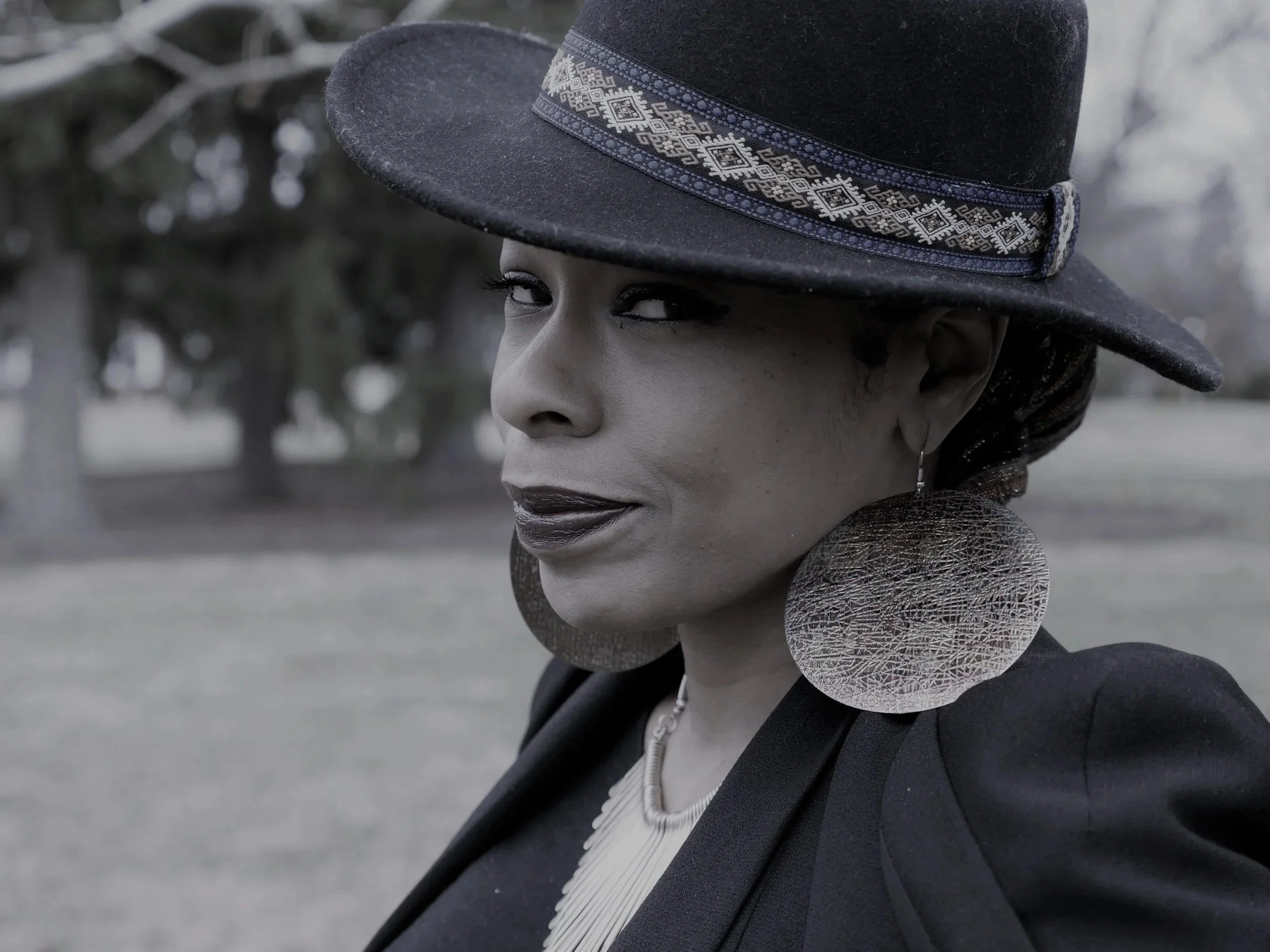 A woman with dark lipstick and jewelry wearing a large black hat with decorative band, outdoors with trees in the background.