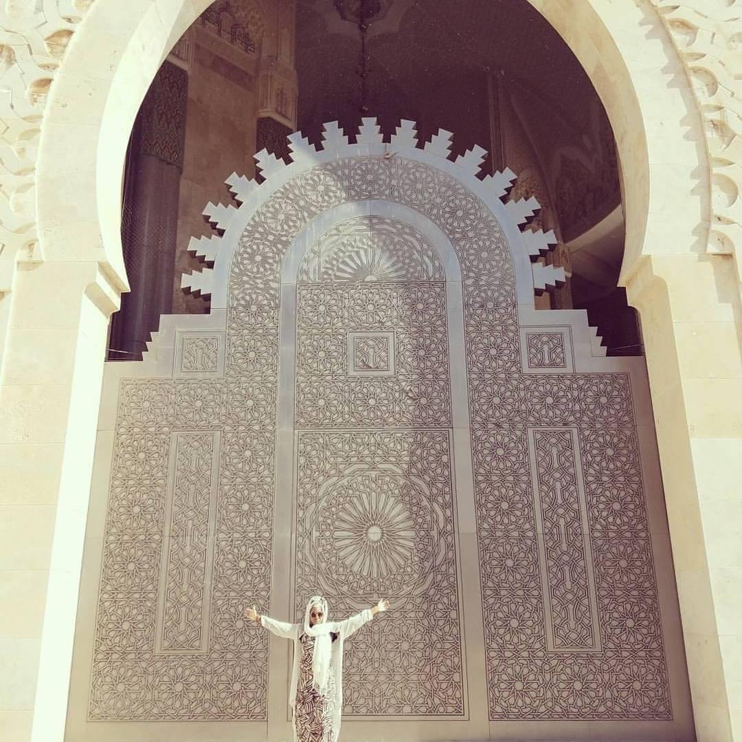 A person standing in front of an ornate, large, arched door in Morocco with intricate geometric patterns, with arms outstretched.